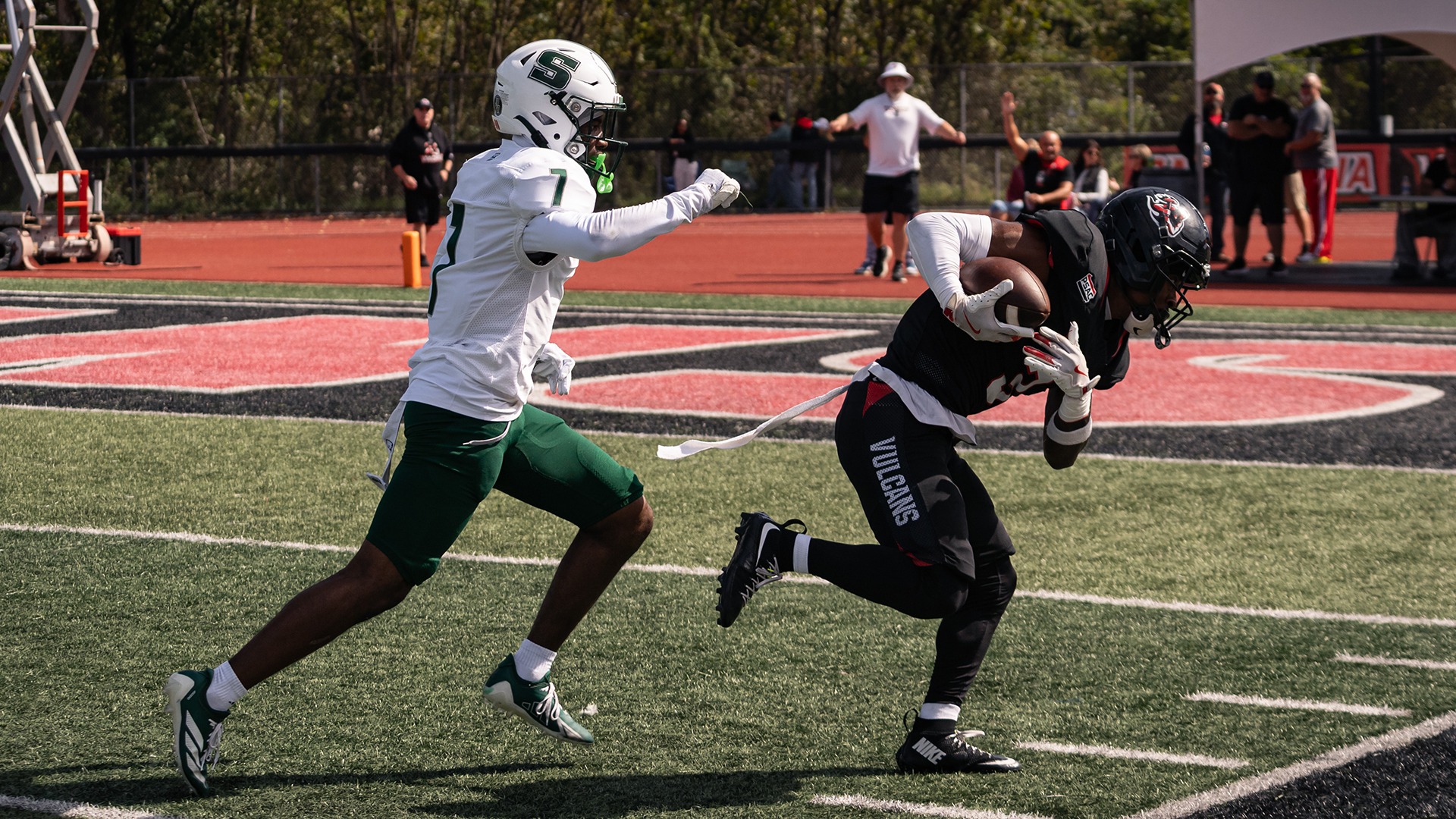 Football player Amire Spencer runs with the football in a dark Vulcans uniform while a Slippery Rock defender in white and green pursues him on the field.
