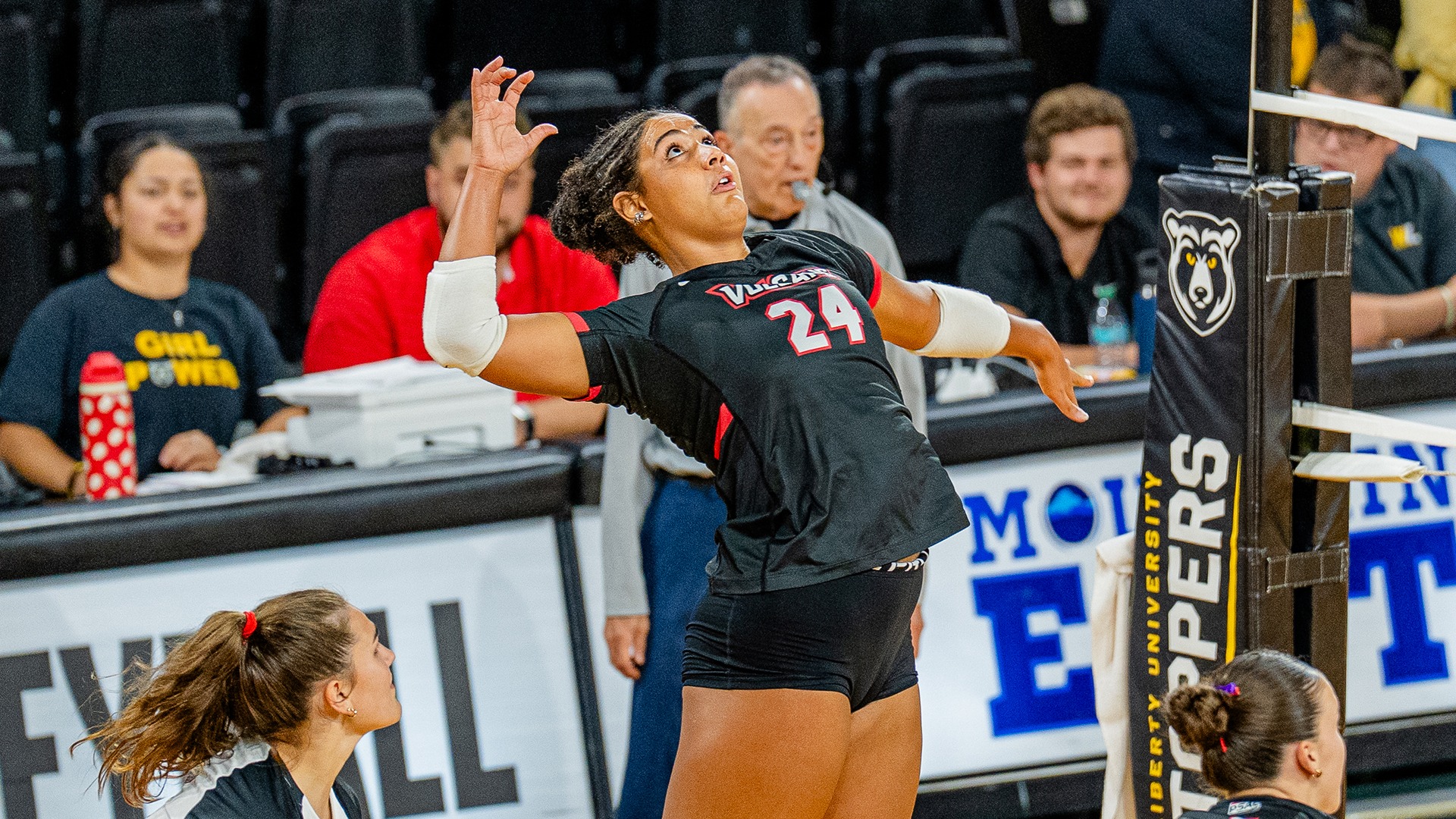 Alayna Jones (#24) arches back mid-air, preparing to hit the volleyball during a match as teammates and spectators look on.