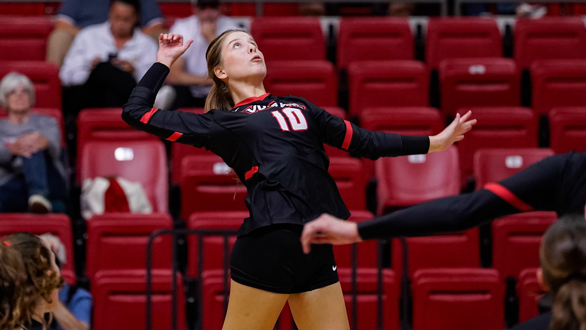 Elise Olenick pulls back her arm to serve a volleyball in a black uniform during a match in an arena with red seating.