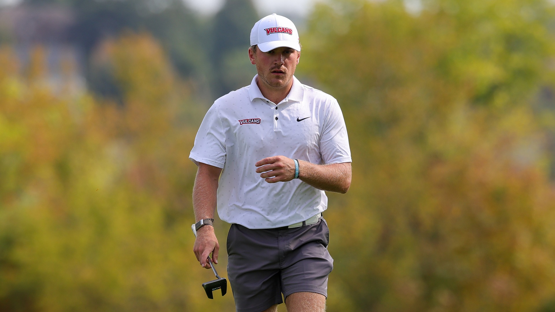 Golfer Anthony DePietress walks across the green carrying a putter while wearing a white Vulcans shirt and white cap.
