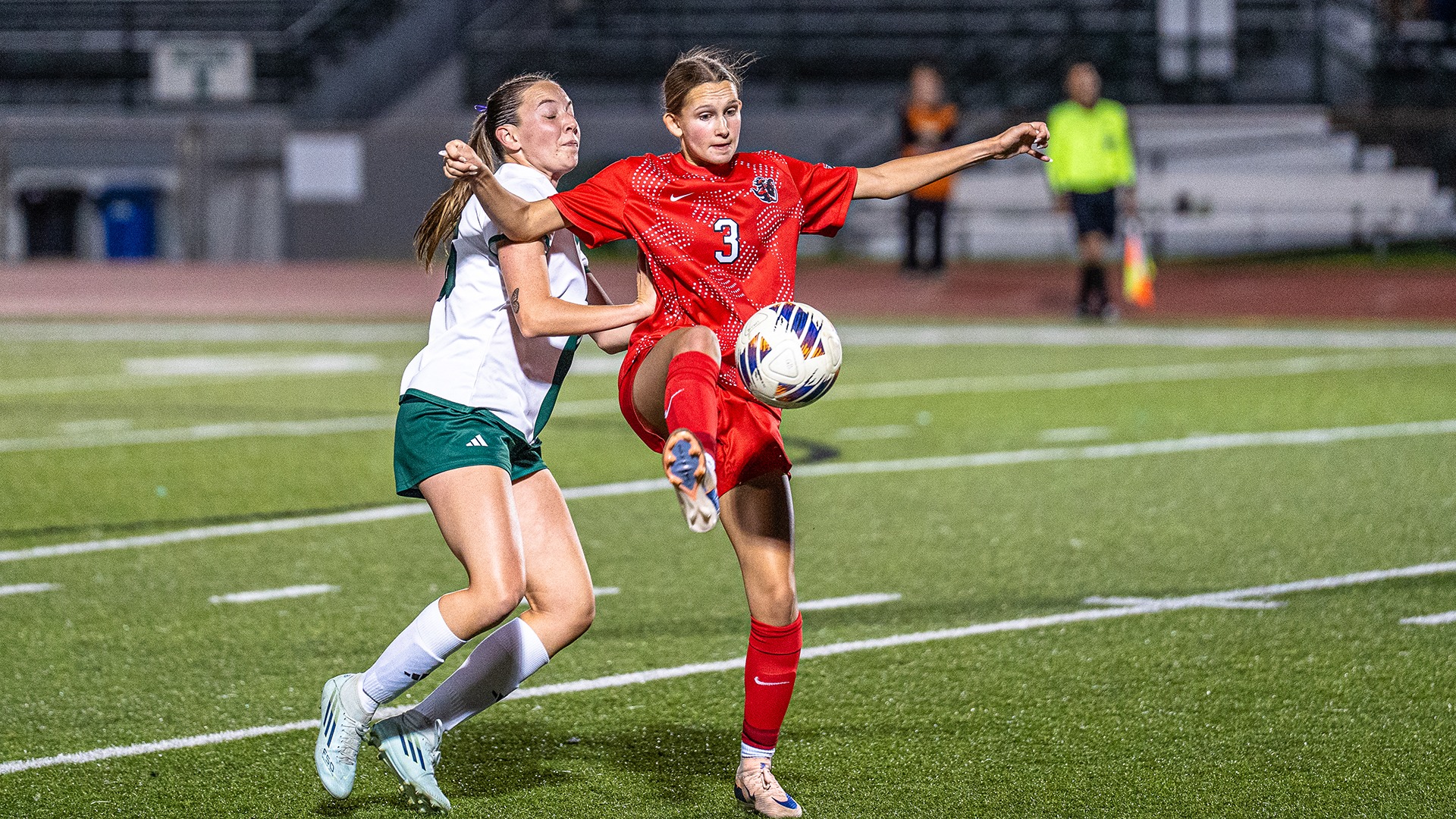 Emma Ziskowski (#3), in a red kit, uses her right foot to control a soccer ball mid-air on a turf field while competing against a opposing player in a white and green jersey.