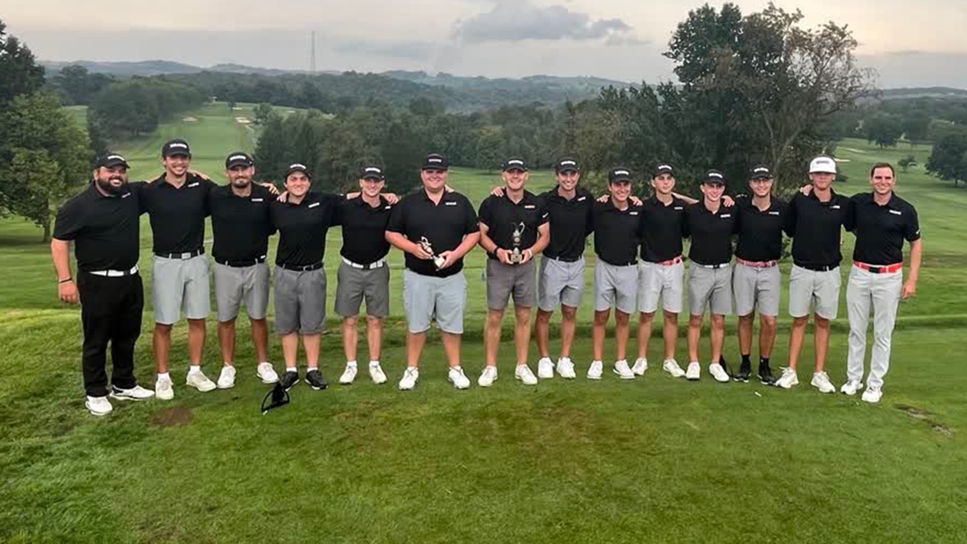 The California men's golf team standing side by side in a line on a grassy hill at Cedarbrook Golf Course, posing with arms around each other's shoulders and holding small trophies.