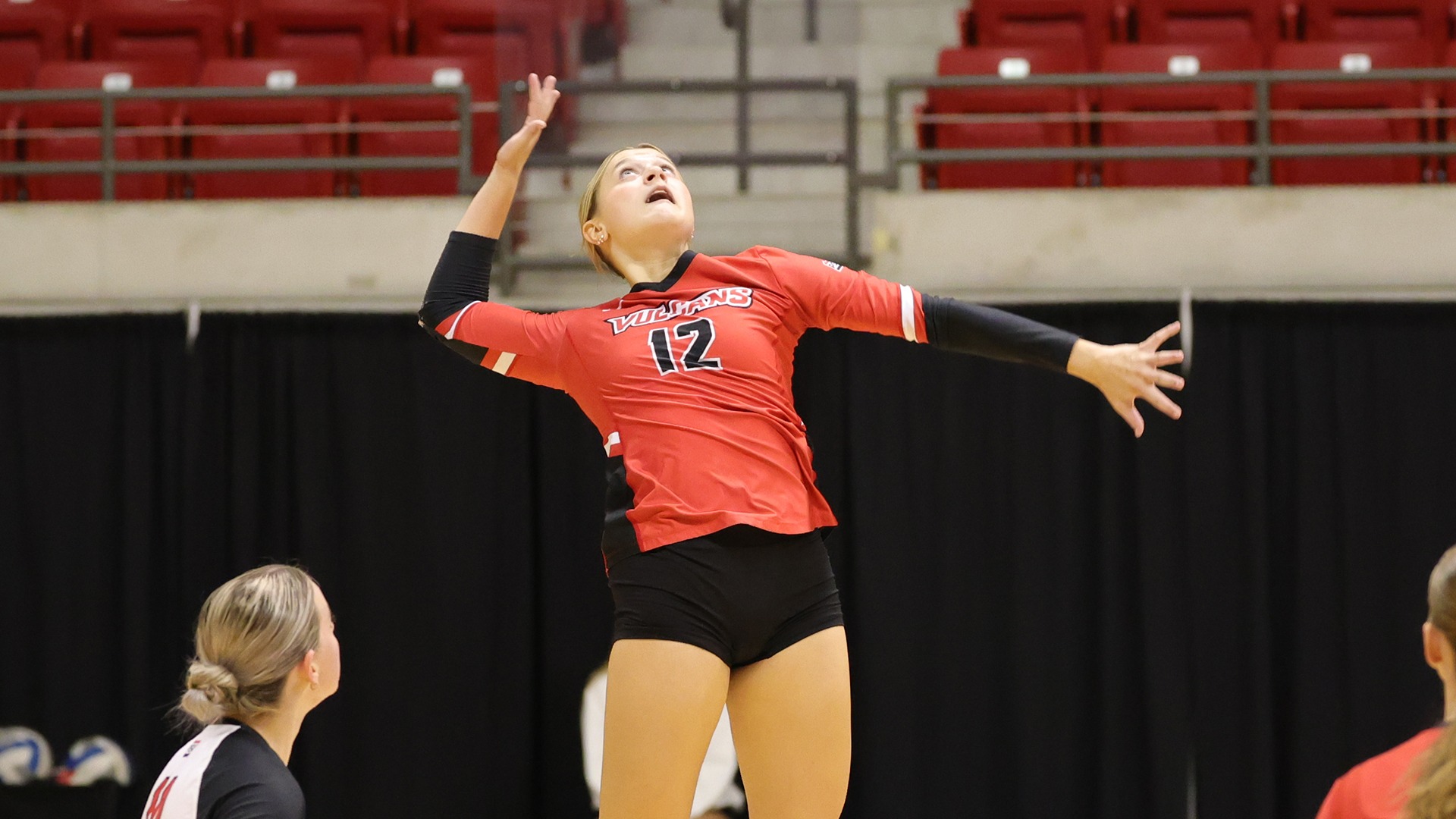 Volleyball player Olivia Weiseman (wearing number 12 in a red jersey and black shorts) leaps high mid-air above the court to hit an incoming ball during a home match. Teammates in red and black uniforms watch from the court and sideline against a background of red arena seating.