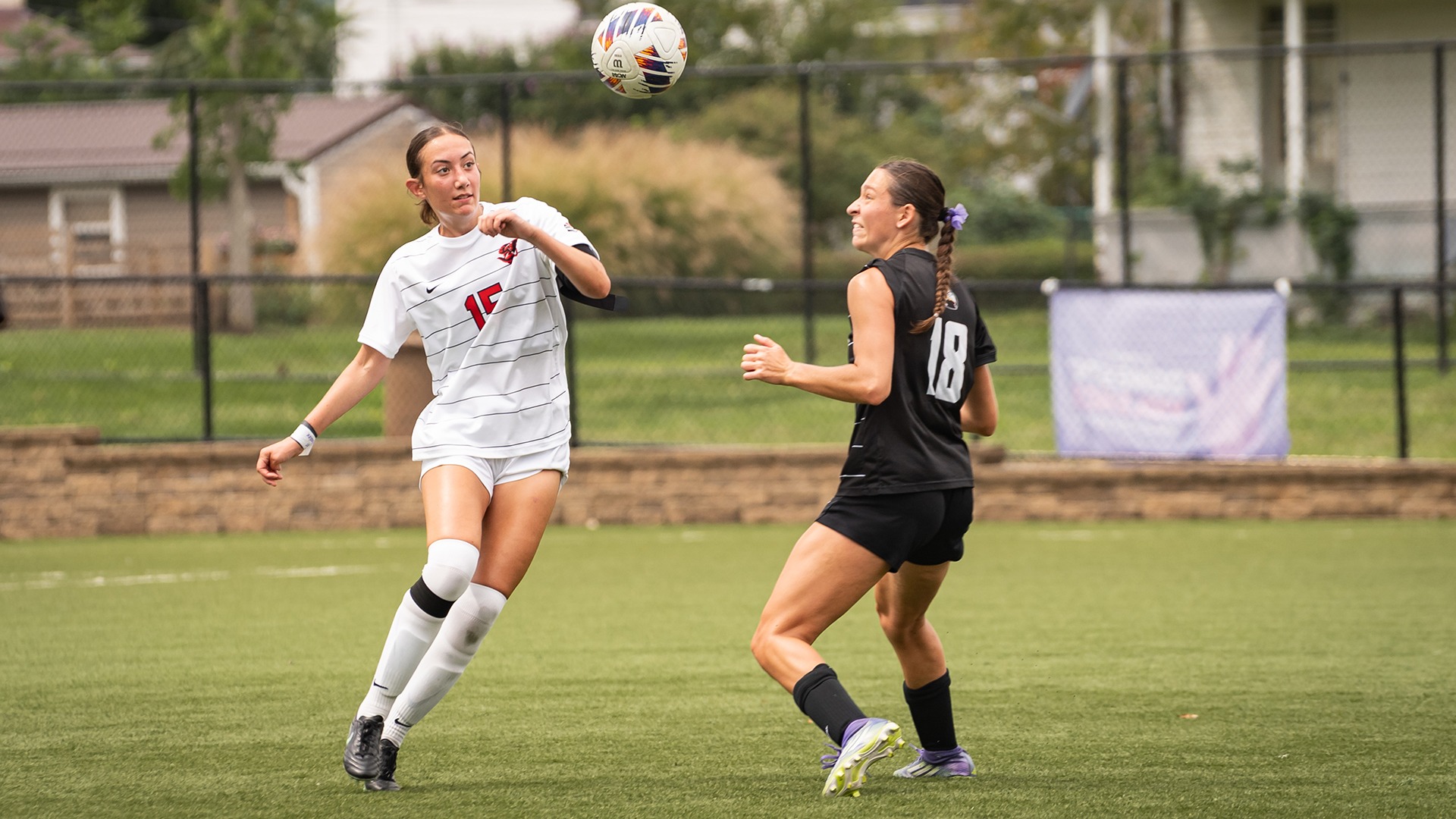 Brooke Barlog, wearing a white soccer jersey with red number 15, tracks a soccer ball floating overhead while a defender in a black jersey (number 18) guards nearby on a turf field.