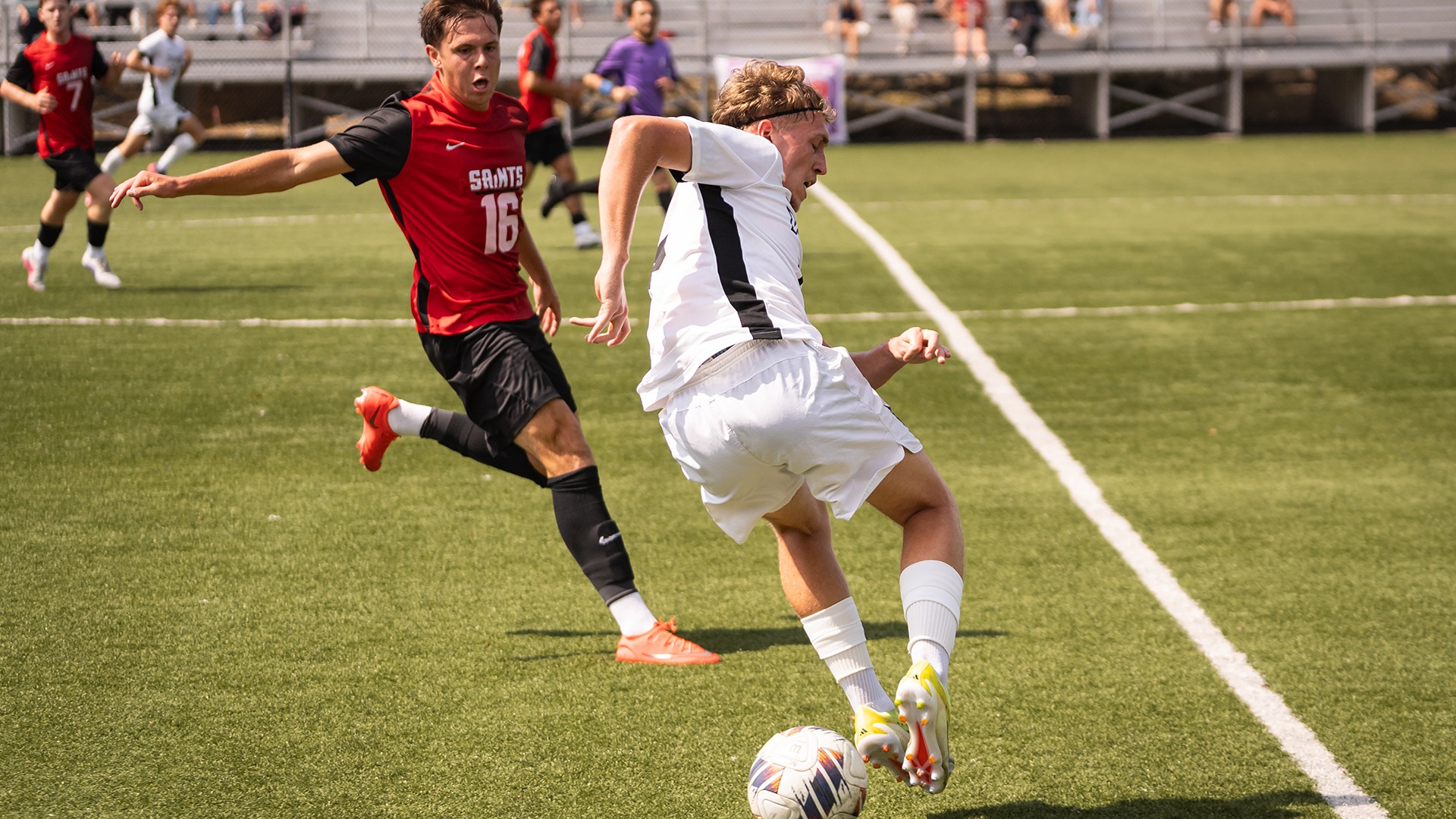 A soccer player in a white uniform maneuvers the ball on a green turf field while being closely pursued by an opponent wearing a red 