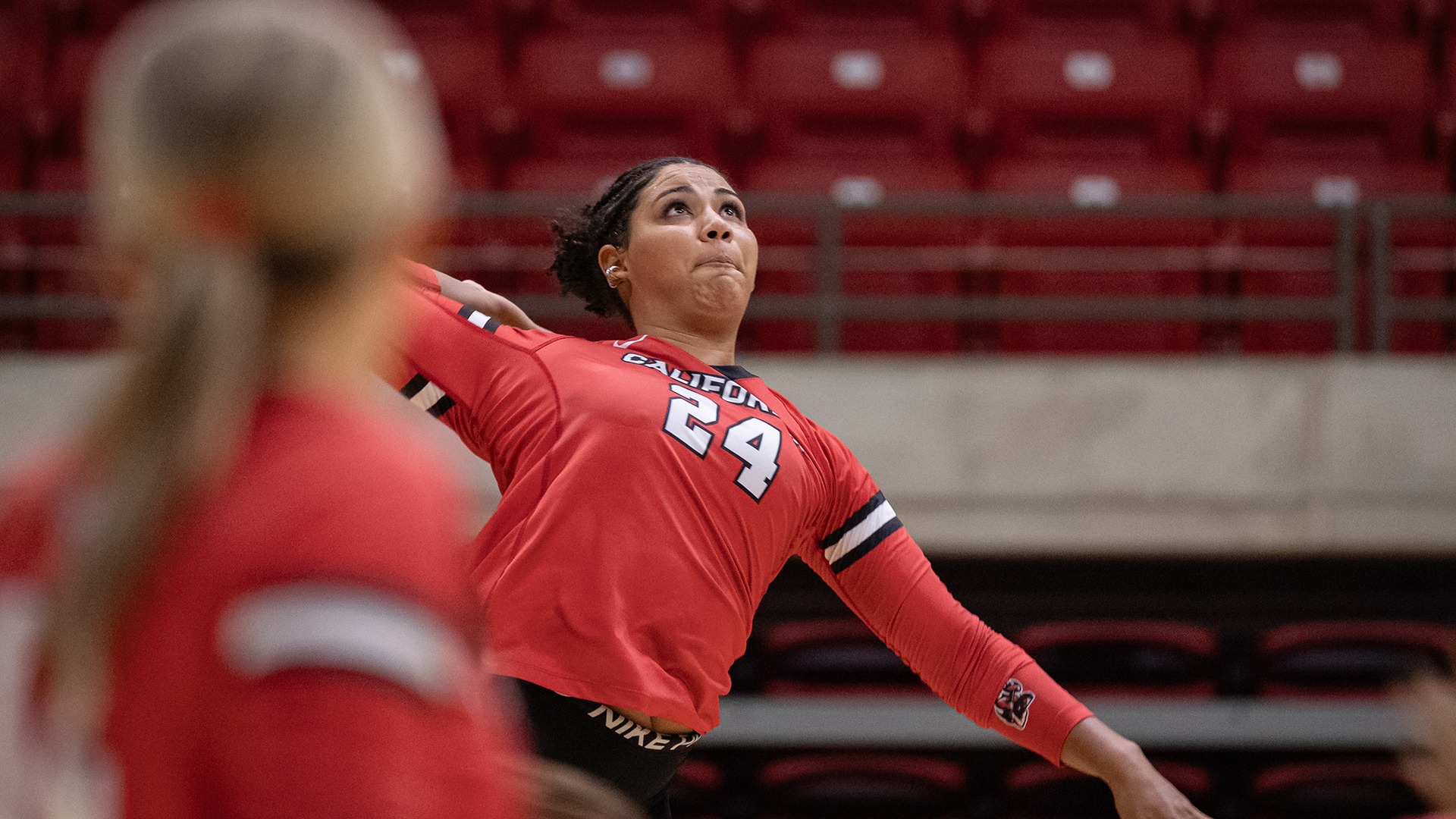 Volleyball player Alayna Jones, wearing a red jersey with the number 24, rears back her arm in mid-action to serve or spike a volleyball during an indoor match in an arena with red seating.