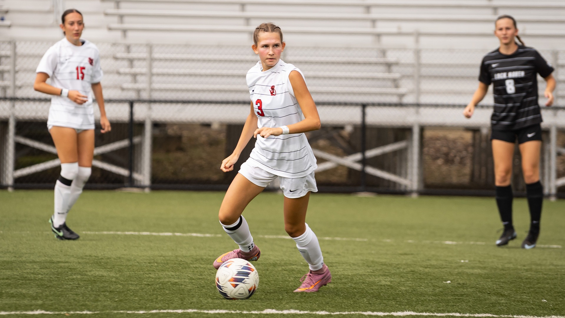 Emma Ziskowski, wearing a white soccer jersey with the number 3, dribbles a soccer ball down the turf field during a match.