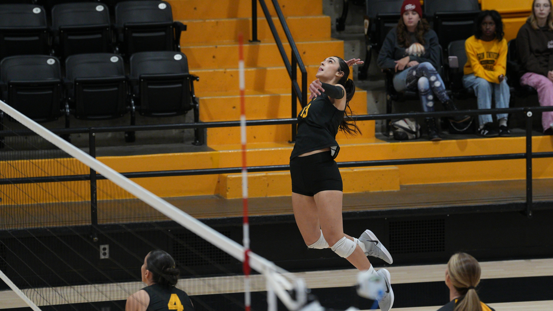 Cameron volleyball player leaps into the air to serve the ball during a match at Aggie Gymnasium, with a teammate positioned near the net.