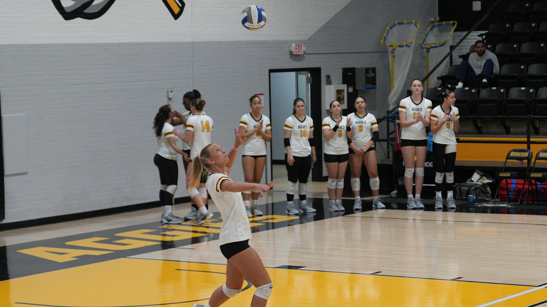 A Cameron University volleyball player prepares to serve during a match at Aggie Gymnasium as teammates watch from the sideline.
