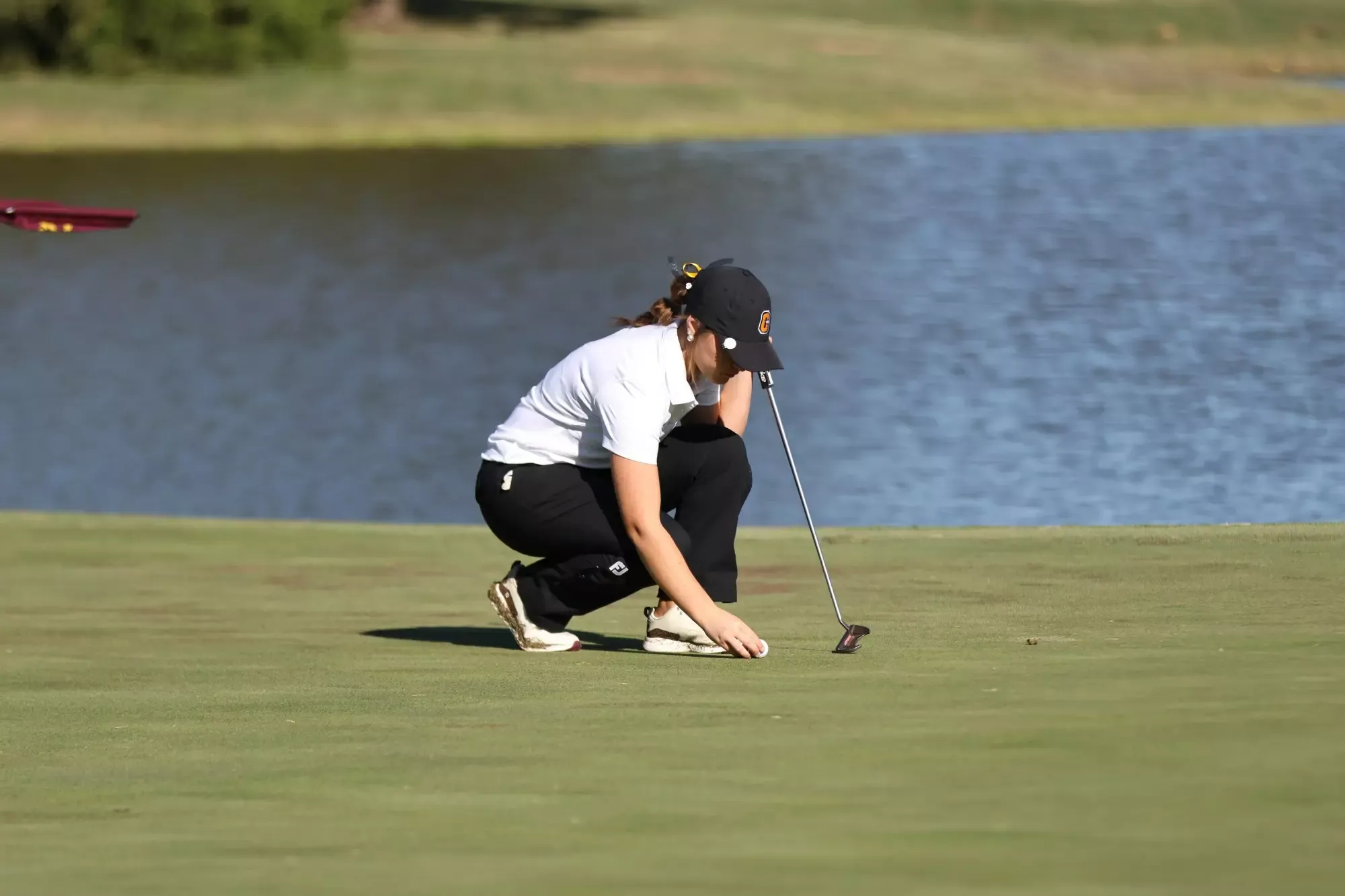 A Cameron University women's golfer crouches on the putting green to position her golf ball while holding a putter, with a pond in the background.