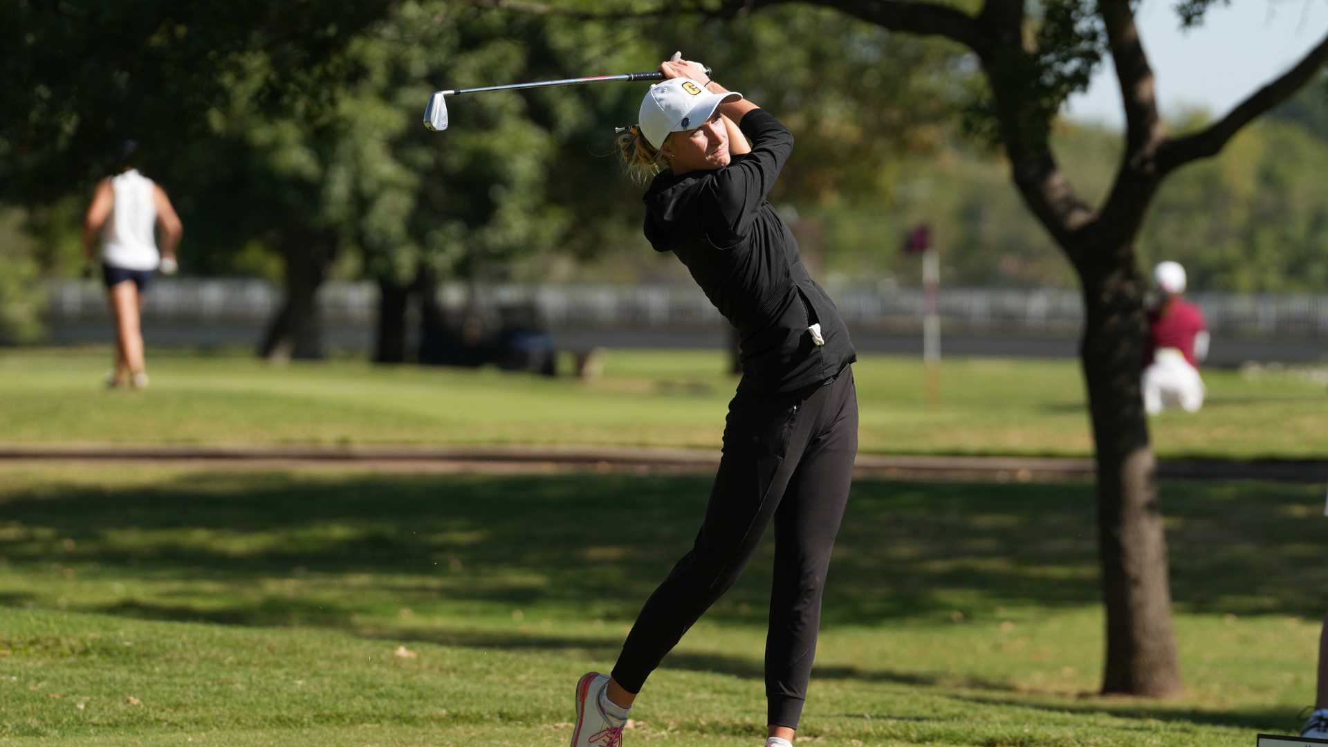 A Cameron University women's golfer wearing a black outfit and white Cameron cap follows through on her swing during a round of golf, with trees and other golfers in the background.