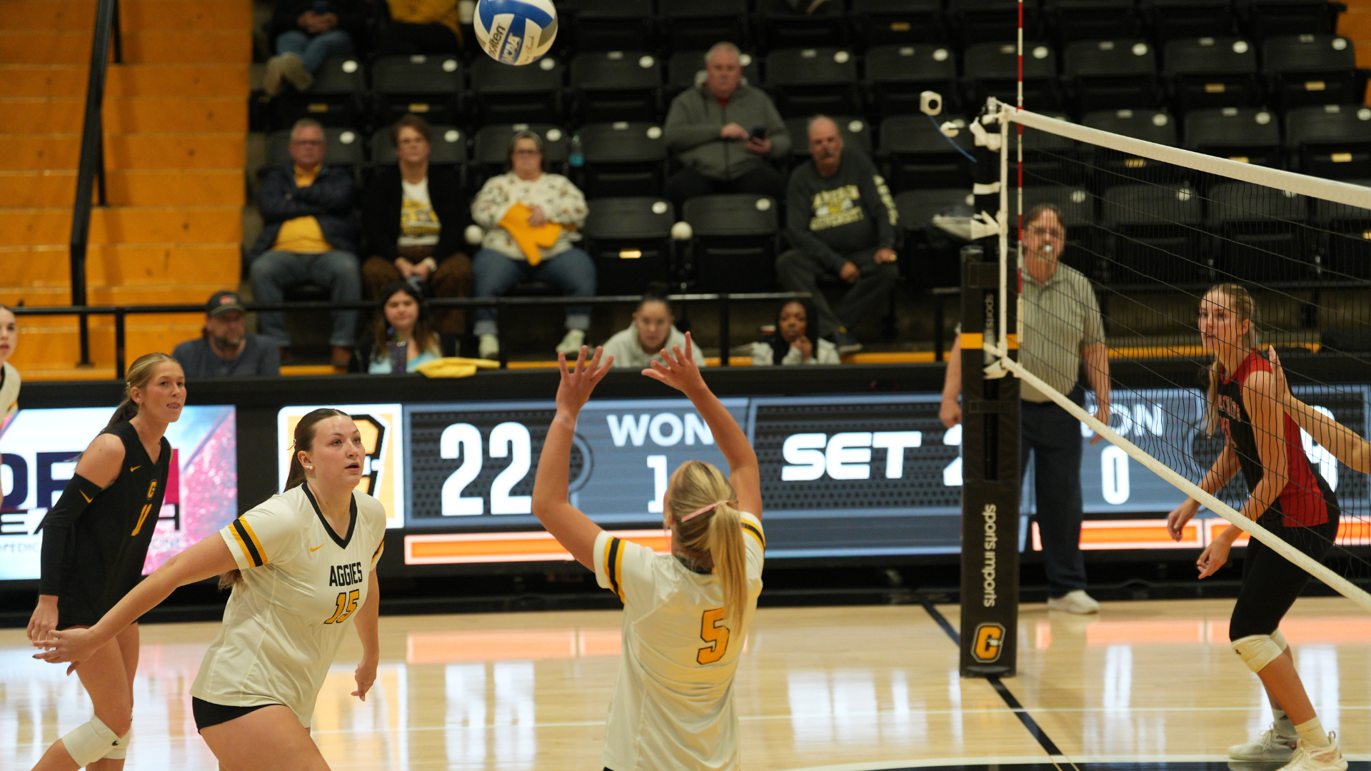 Cameron University volleyball player No. 5 prepares to set the ball as teammates No. 15 and No. 1 position themselves during a match at Aggie Gym. An opposing player stands near the net while spectators watch from the stands.