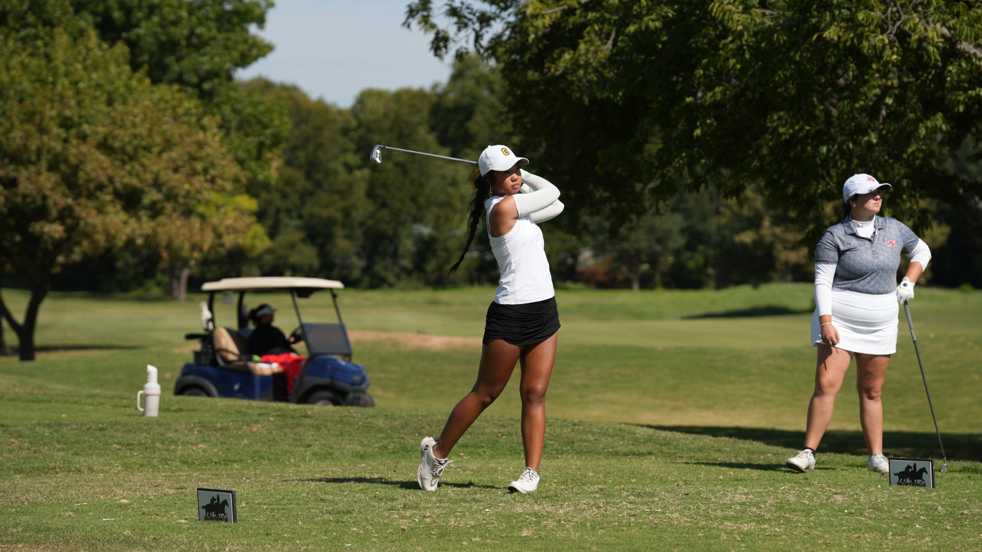 Cameron University women's golfer follows through on an iron shot on a sunny golf course, wearing a white Cameron cap, white sleeveless top, and black skirt. Another golfer stands nearby watching, with a golf cart and trees in the background.
