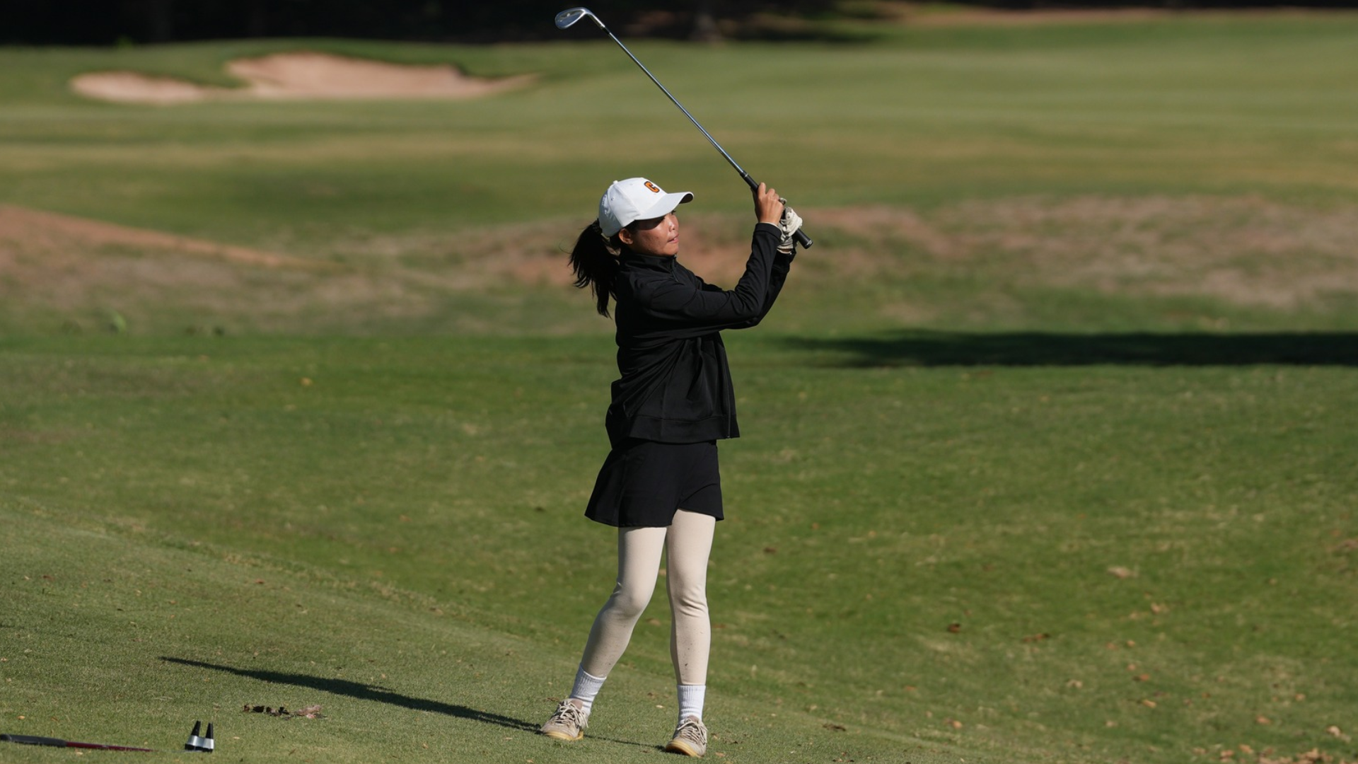 Cameron University women's golfer follows through on an iron shot from the fairway, wearing a white Cameron cap, black jacket, black skirt, and white leggings on a sunny golf course.