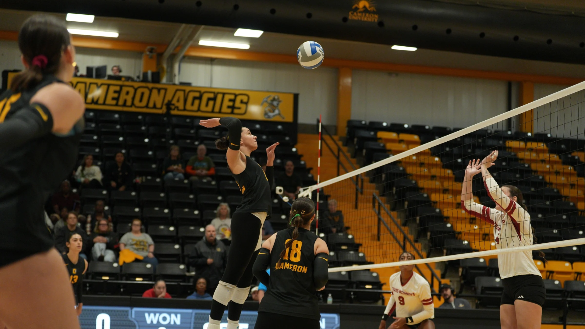 Cameron University volleyball player prepares to attack the ball at the net during a home match at Aggie Gymnasium, while a Texas Woman’s University defender jumps with both hands raised to block. Cameron teammates look on as the play unfolds.