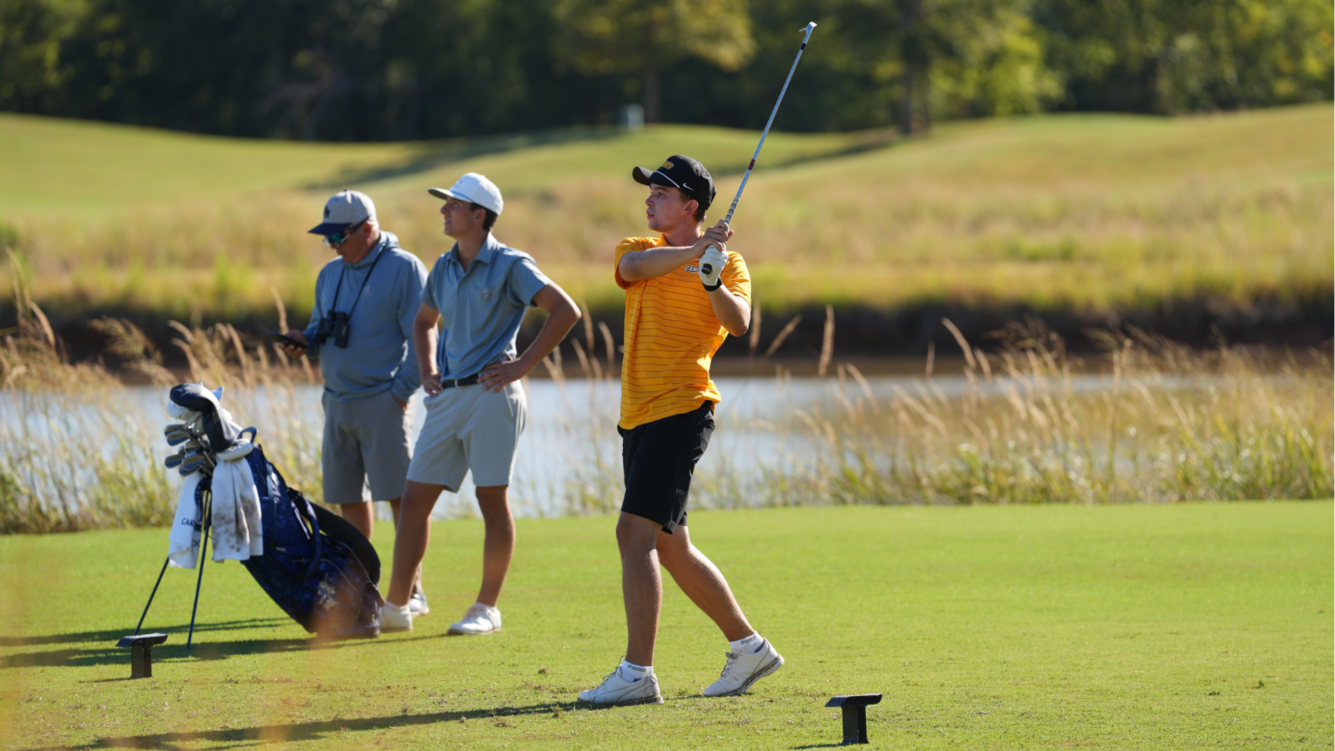 Cameron men’s golfer watches his shot after a swing on the course, with two people and a golf bag in the background near a water hazard.