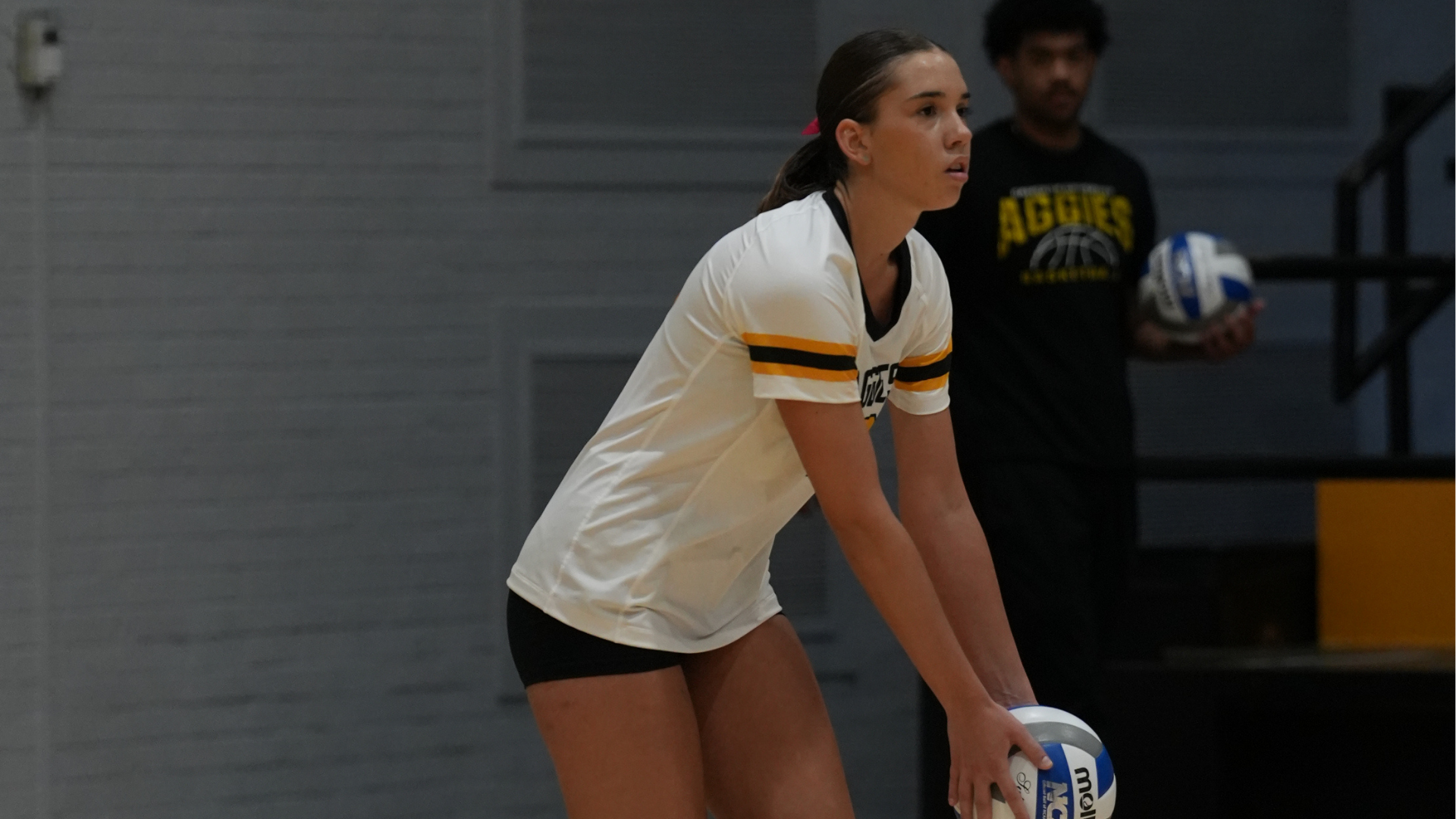Cameron volleyball player in a white, black and gold uniform prepares to serve during a match, holding a volleyball while focused on the court.