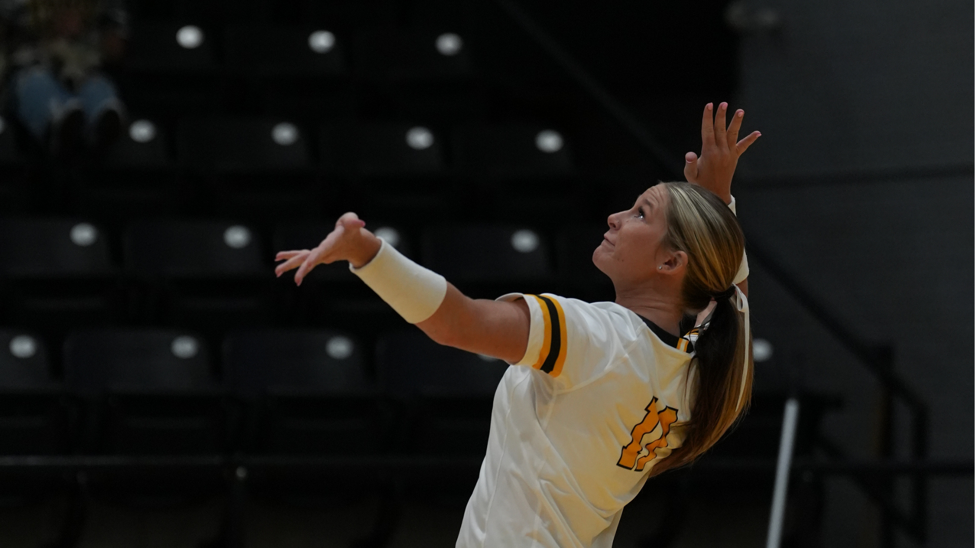 Cameron volleyball player in a white, black and gold uniform reaches up and prepares to serve during a match.