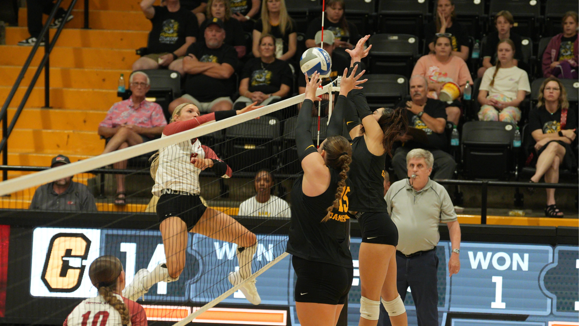 Cameron volleyball players jump to block an attack from Southern Nazarene during a match inside Aggie Gym, with a large crowd of fans visible in the stands behind the court.