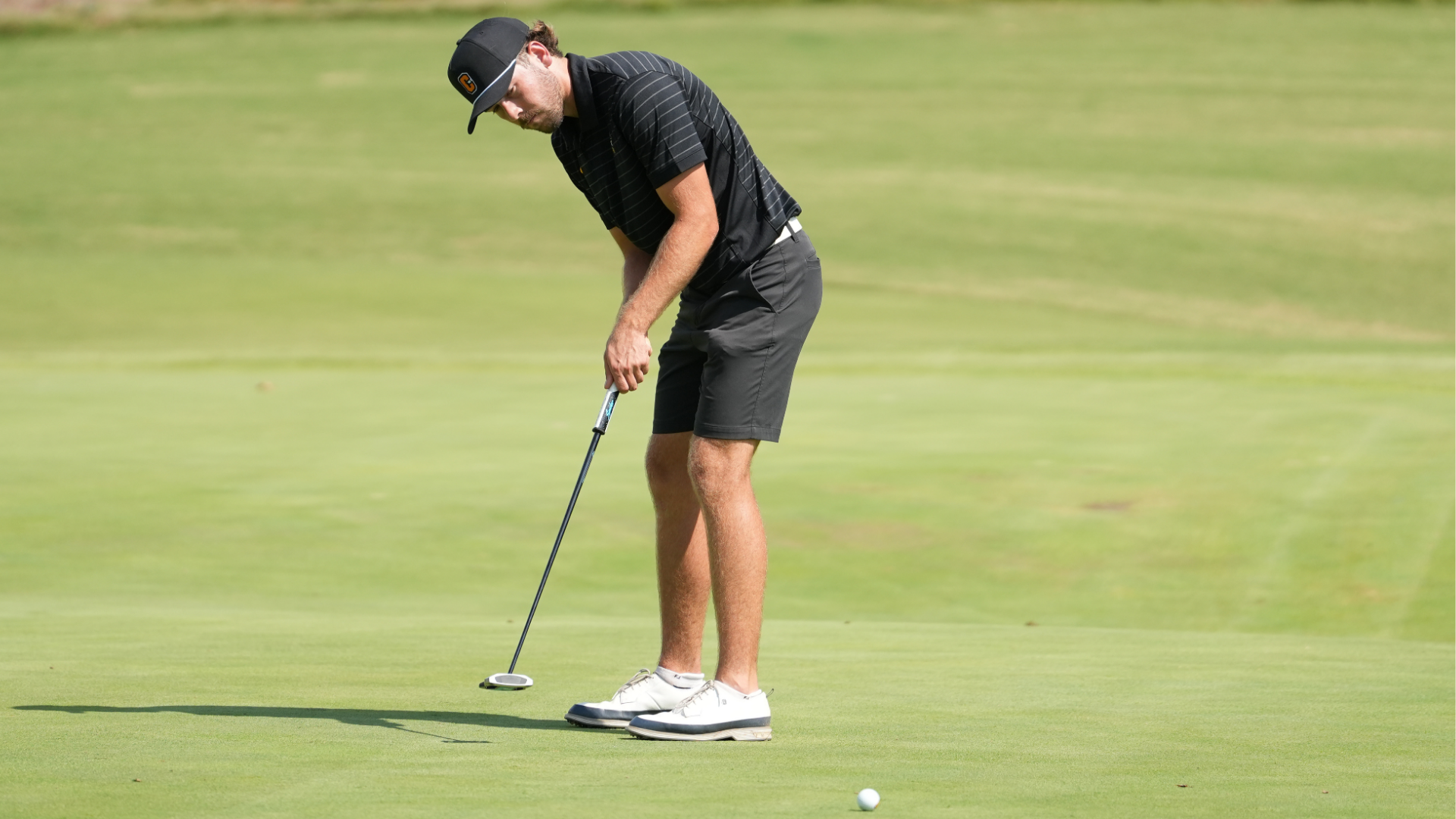 Cameron University men’s golfer prepares to putt on a green during competition, wearing a black Cameron cap and black golf attire.