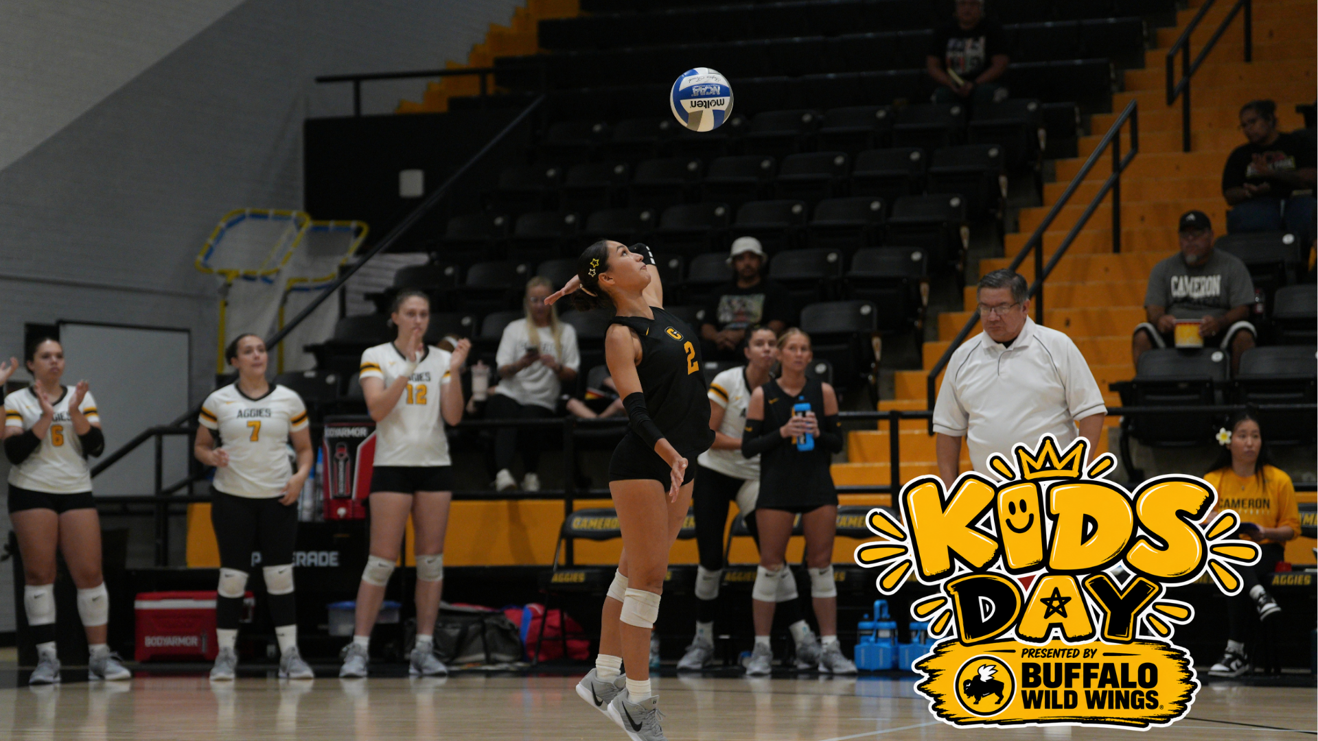 Cameron volleyball player serves the ball during a match inside Aggie Gym, with teammates watching from the sideline with Kids Day presented by Buffalo Wild Wings logo