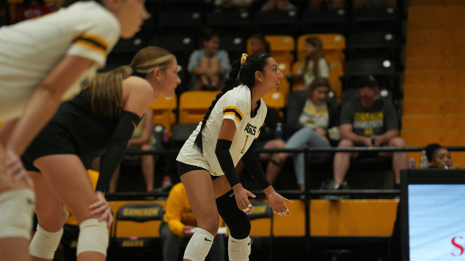 Cameron University volleyball players stand in a defensive ready position on the court during a match, with a player in a white Aggies jersey in focus and teammates positioned beside her.