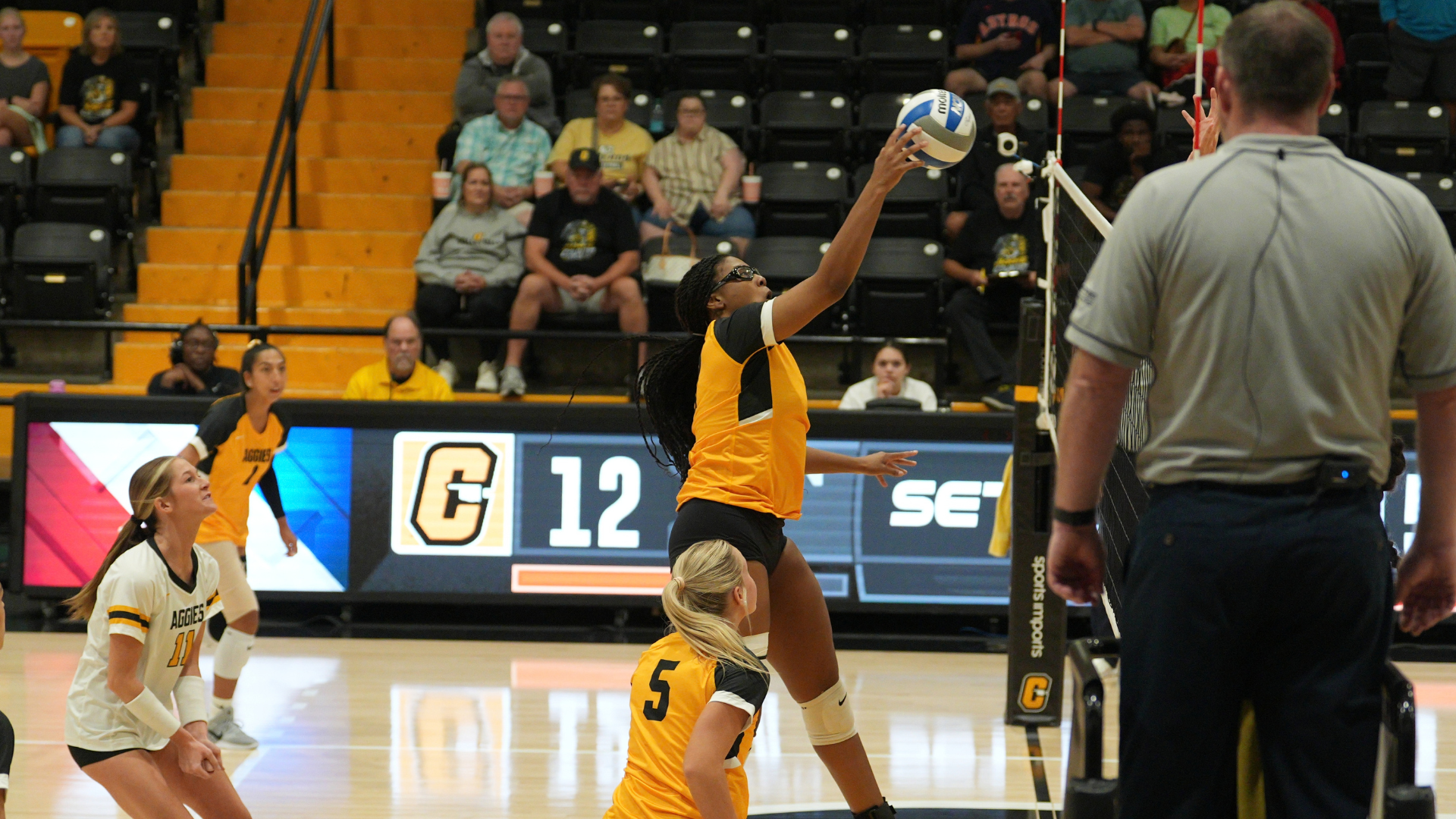Cameron University volleyball player reaches up to attack the ball at the net during a match as teammates look on from the court.
