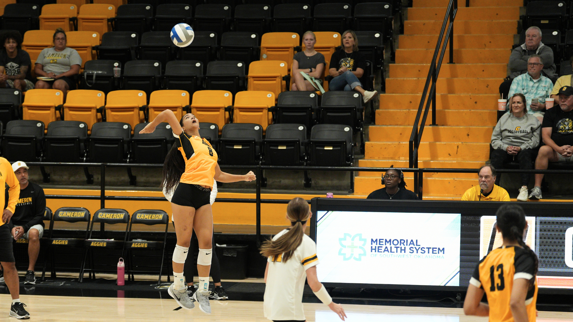 Cameron volleyball player serves the ball during a match inside Aggie Gym, with teammates, spectators, and the scorer’s table visible in the background.