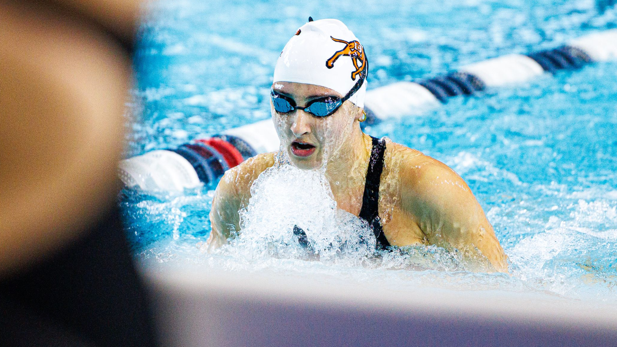 A Campbell swimmer wearing a white swim cap and goggles powers through a breaststroke lap, water splashing around her as she approaches the lane divider.