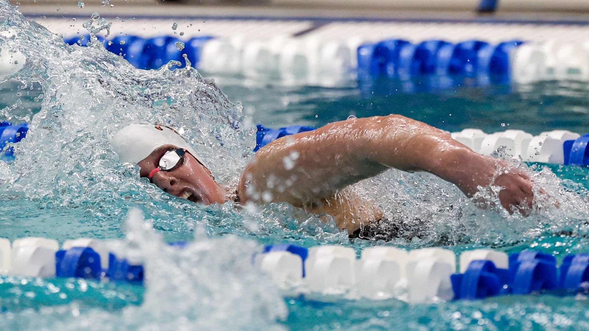 Campbell's Katarina Gagnon, wearing a white cap and mirrored goggles, competes in the 500 freestyle in a lane-marked pool, with water splashing around their arm during a stroke at the 2026 CAA Championships.
