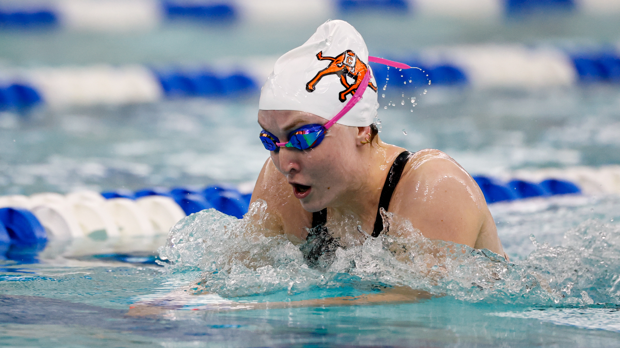 Arabella Butera, wearing a white swim cap with an orange Campbell Fighting Camels logo and pink-strapped goggles performing the breaststroke in the 2026 CAA Championships, water splashing around her as blue and white lane lines blur in the background.