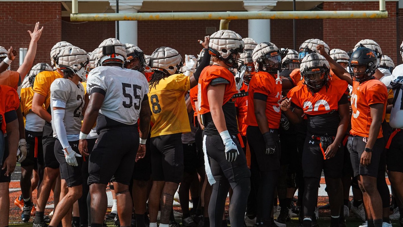 A group of Campbell football players in practice uniforms huddle together on the field at Barker-Lane Stadium, wearing helmets and raising their hands in a team chant near the goalpost.