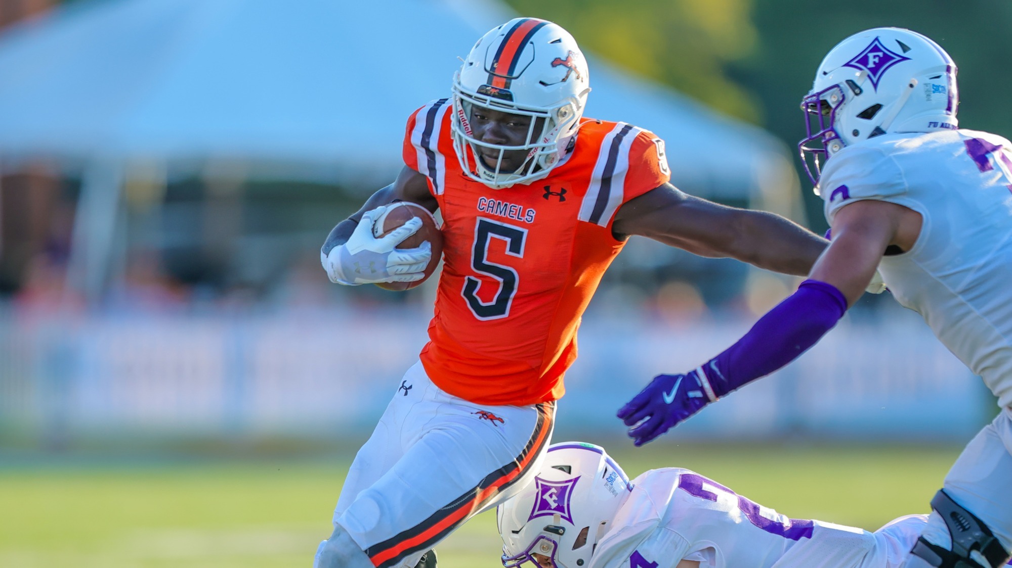 Campbell's JJ Cowan, in an orange jersey (number 5), runs with the ball, fending off a Furman defender in a white and purple uniform while another defender attempts a tackle from below.