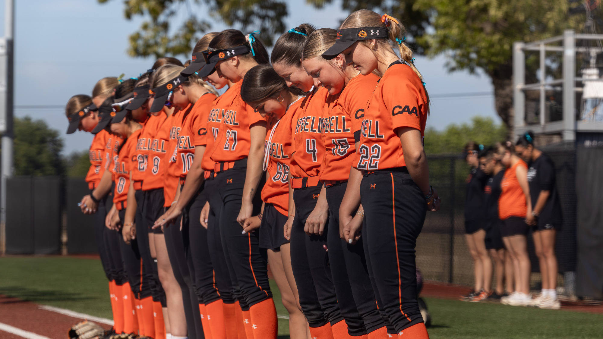 Campbell softball players stand in a line along the foul line with heads bowed during a pregame moment, wearing orange jerseys and black pants on a sunny day.