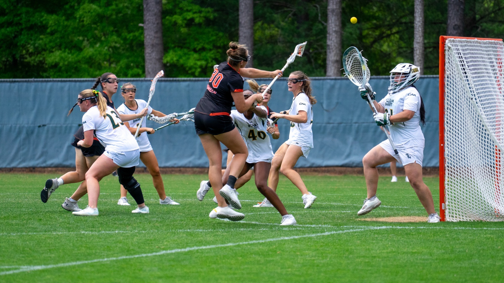 #30 Drew Watkins in a black jersey leaps to shoot at the goal, surrounded by William & Mary defenders in white. The ball is mid-air traveling above the goalkeeper's head towards the goal.