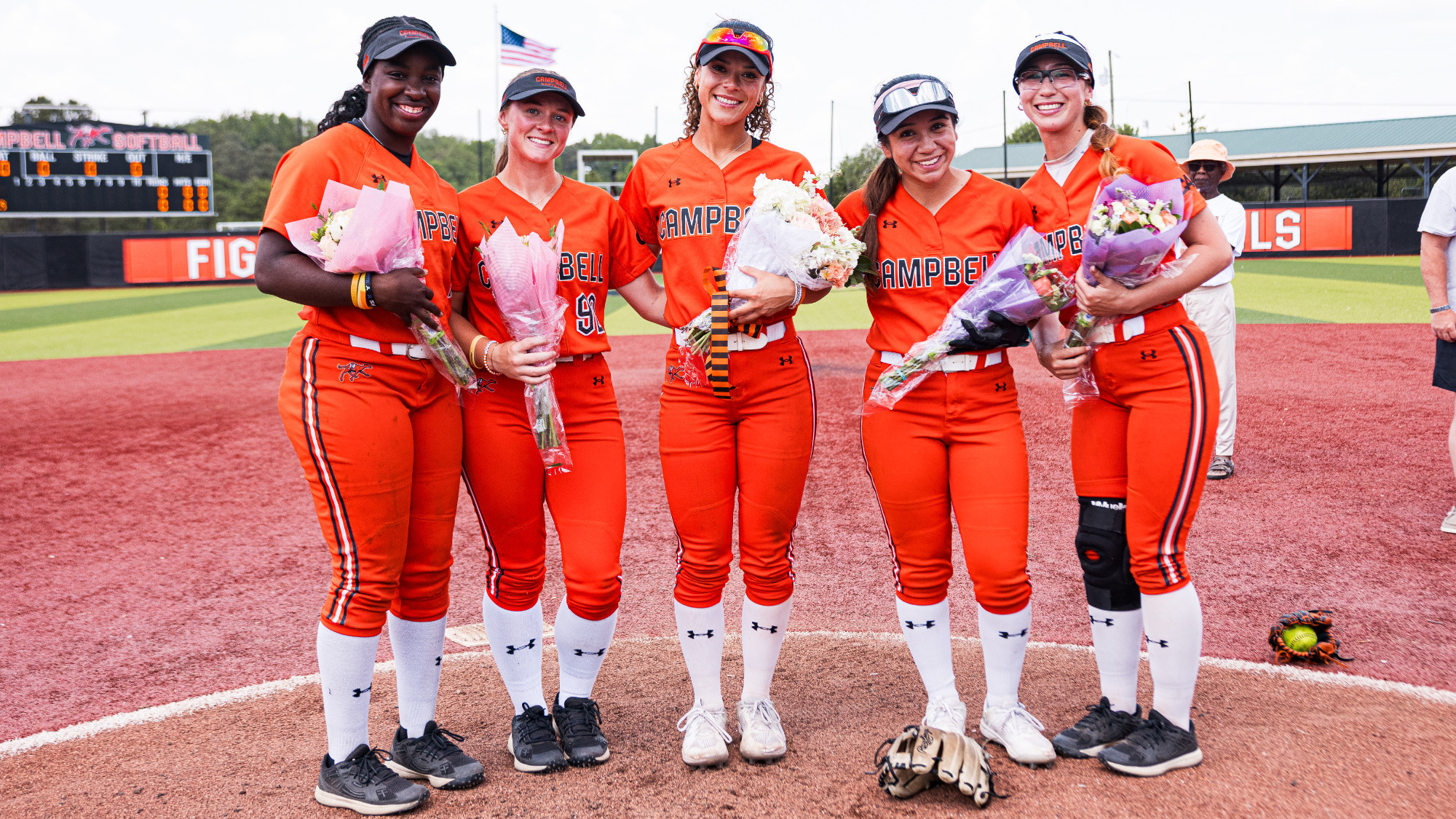 Alyssa Armijo, Amber DeSena, Megan Leon, Charlie Montgomery, and Allyiah Swiney stand together at Amanda Littlejohn Stadium with flowers, celebrating senior day