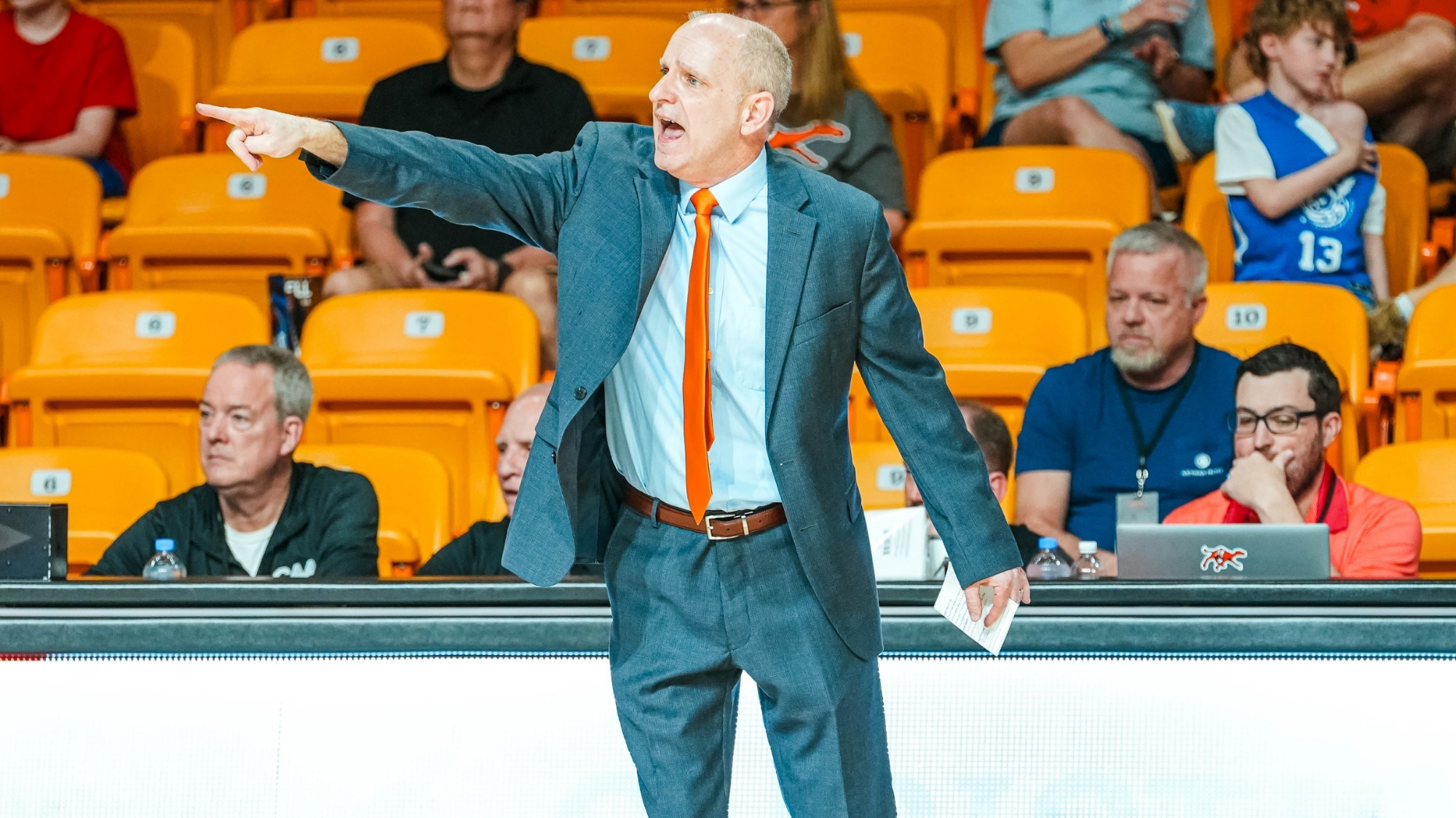 Campbell women's basketball head coach Ronny Fisher in a suit pointing with his right hand in front of the stands.