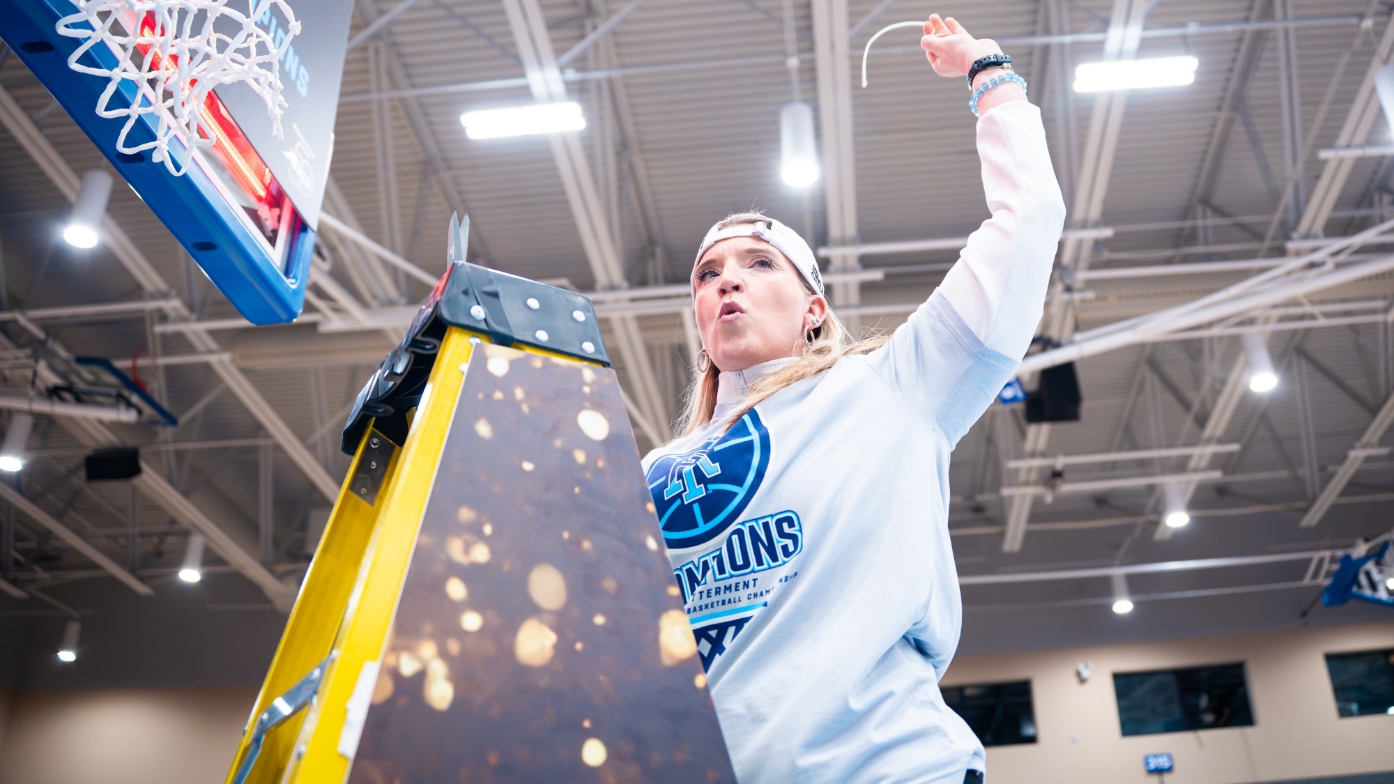 New Campbell head women's basketball coach Ali Jaques, pictured in a white Rhode Island championship shirt and backward cap stands on a ladder beside a basketball hoop, cutting down the net in a brightly lit gym in celebration of the 2026 Atlantic 10 Tournament Championship.