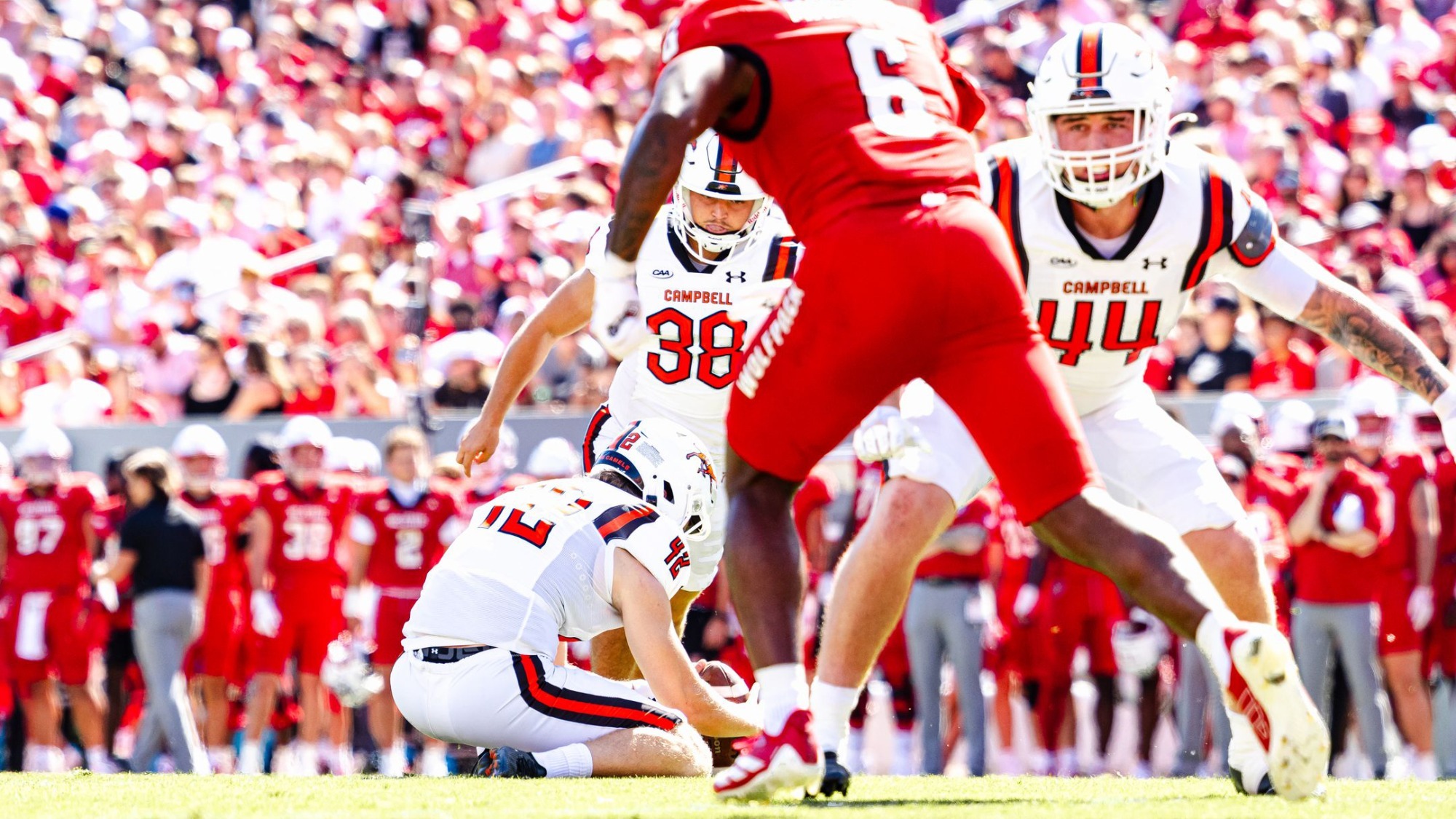 A college football game in progress, viewed from field level. A player in a white Campbell uniform kneels to hold the football for a kick while teammates in white prepare around him. Opposing players in bright red uniforms move across the foreground, partially blocking the view. A crowded stadium filled with fans dressed mostly in red blurs into the background under bright daylight.