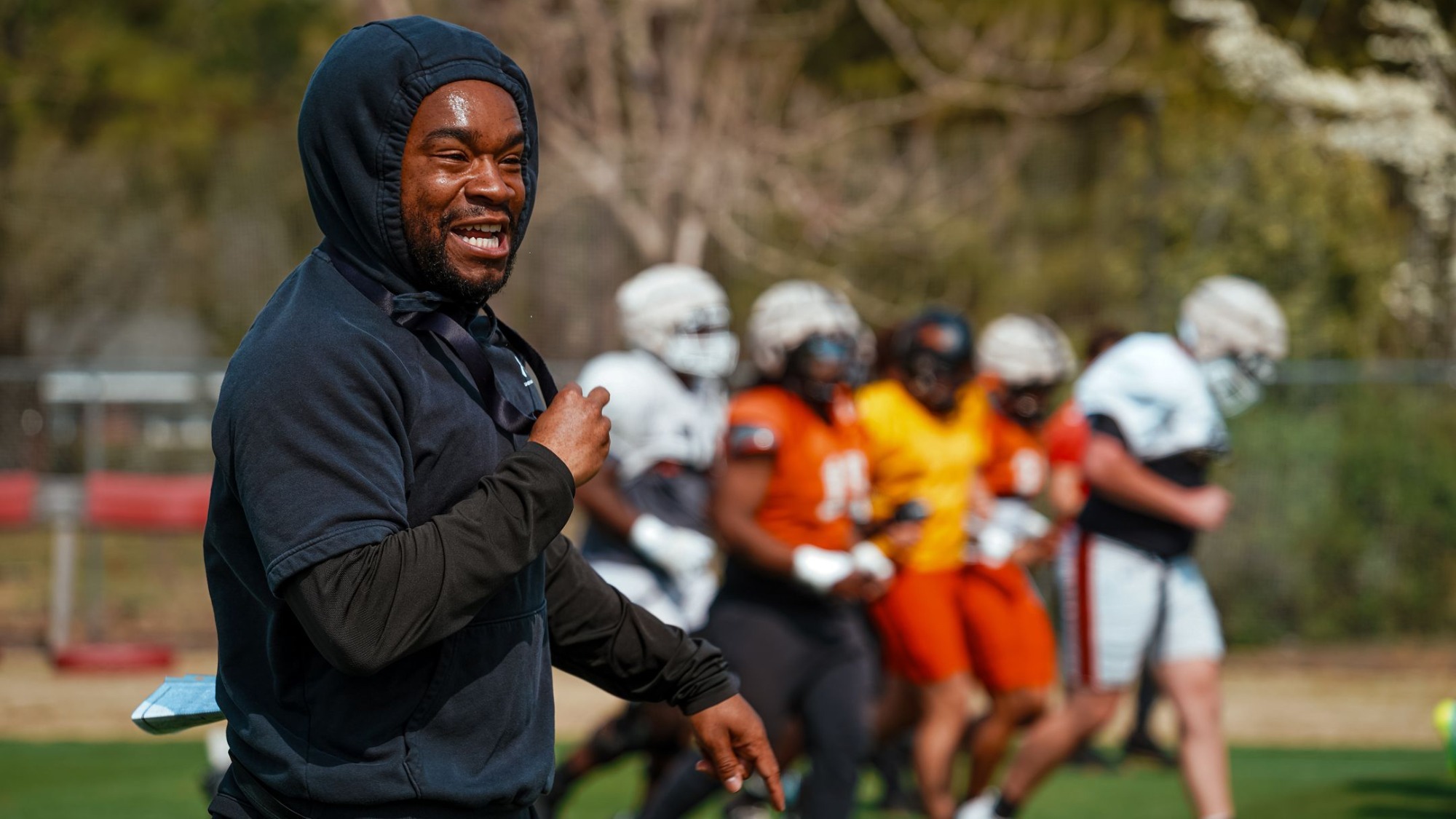 A smiling Larry Dace III in a black hoodie gestures during an outdoor football practice, with players in helmets and colorful jerseys blurred in the background.