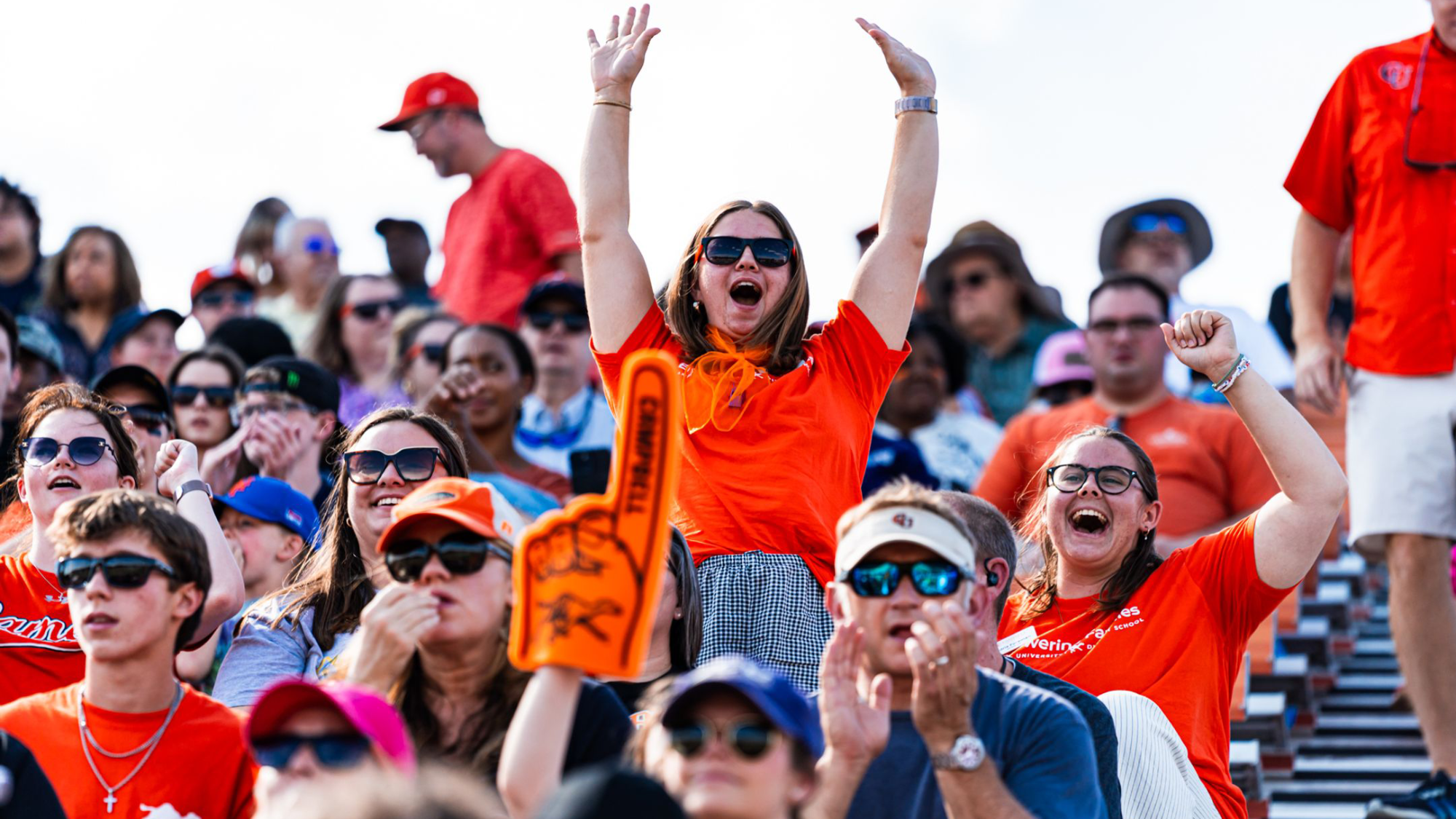 A crowd of fans in bright orange shirts cheers from the Barker-Lane Stadium stands during a Campbell football game. At the center, a woman wearing sunglasses raises both arms overhead and shouts in celebration, while another fan nearby pumps a fist and smiles. Other spectators clap, watch the action, and hold an orange foam finger, creating an energetic game-day atmosphere.