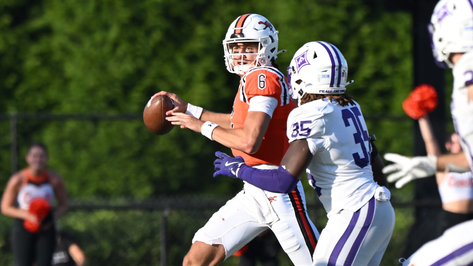 Campbell's quarterback, Kamden Sixkiller, in an orange jersey and white helmet drops back to pass while being pressured by a defender in a white-and-purple uniform. The quarterback cradles the football and looks downfield as the defender closes in from his right side. Additional players and cheerleaders appear blurred in the background, with green trees behind the field on a sunny day.