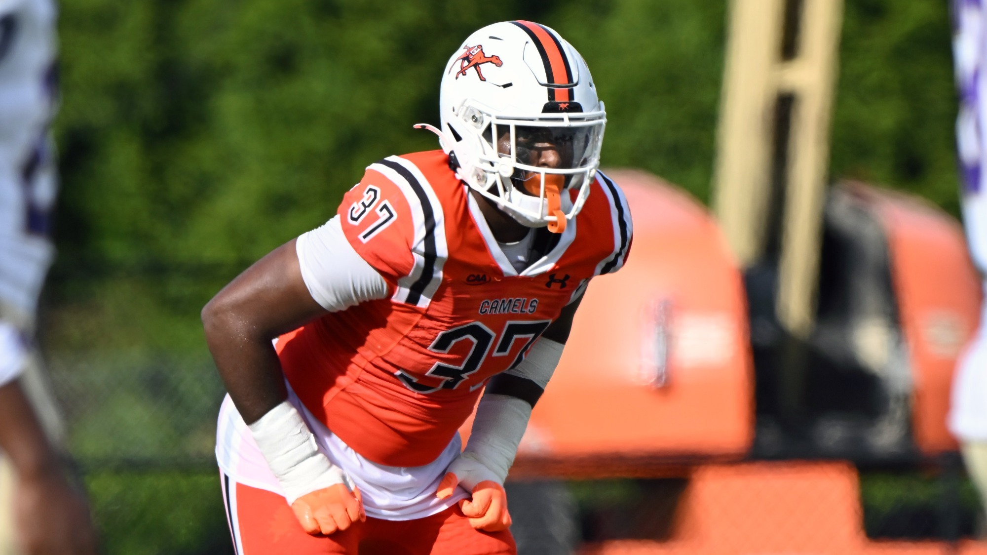 **Alt text:**  DaVonyae Pettis, a Campbell football player wearing an orange jersey (#37) and white helmet crouches in a ready defensive stance before the snap during a daytime game. The athlete wears white arm sleeves and orange gloves, with a blurred football field and sideline equipment in the background.