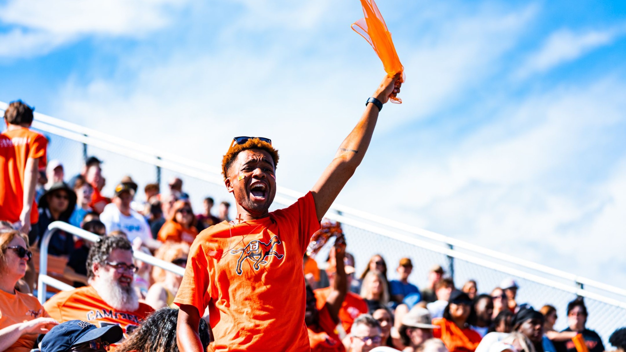 **Alt text:** A Campbell Athletics fan wearing a bright orange t-shirt stands in the Barker-Lane Stadium bleachers with one arm raised, waving an orange rally towel. The fan appears to be shouting enthusiastically, while a crowd of spectators dressed mostly in orange fills the background under a blue sky with scattered clouds.