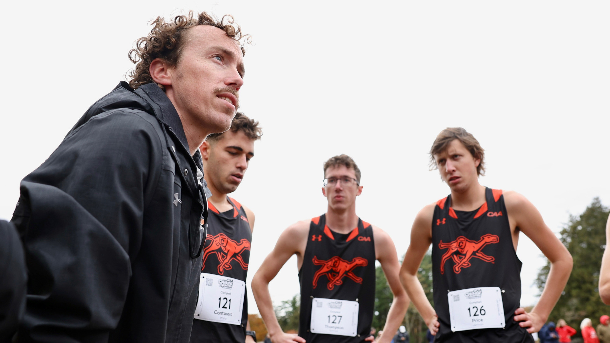 Head coach Evan Darm stands with three Campbell cross-country runners stand outdoors during a race, wearing black and orange uniforms with race bibs.