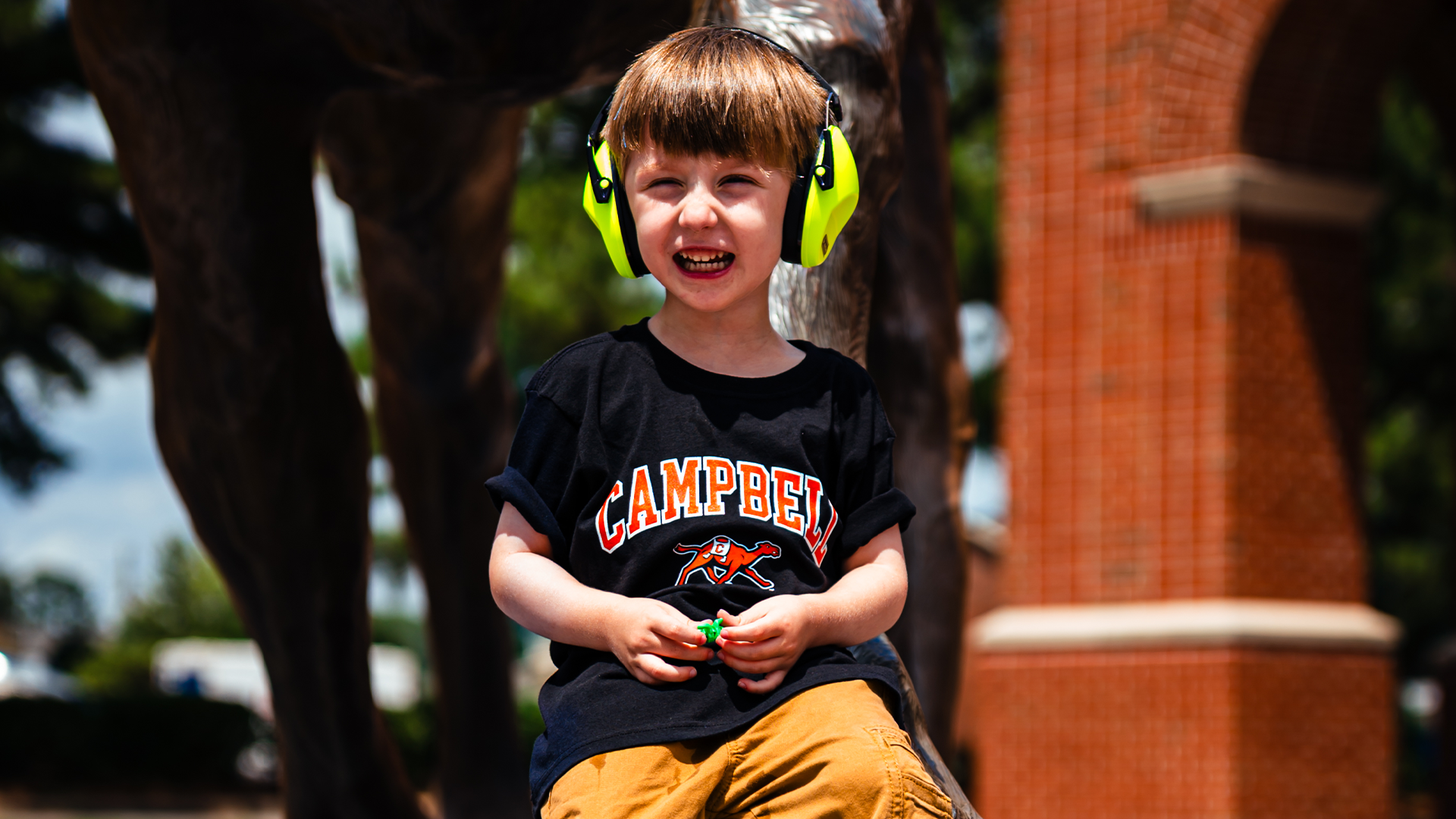 A young child wearing a Campbell Athletics T-shirt, khaki pants, colorful sneakers, and bright green hearing protection sits in front of Campbell University’s camel statue on campus, smiling at the camera.