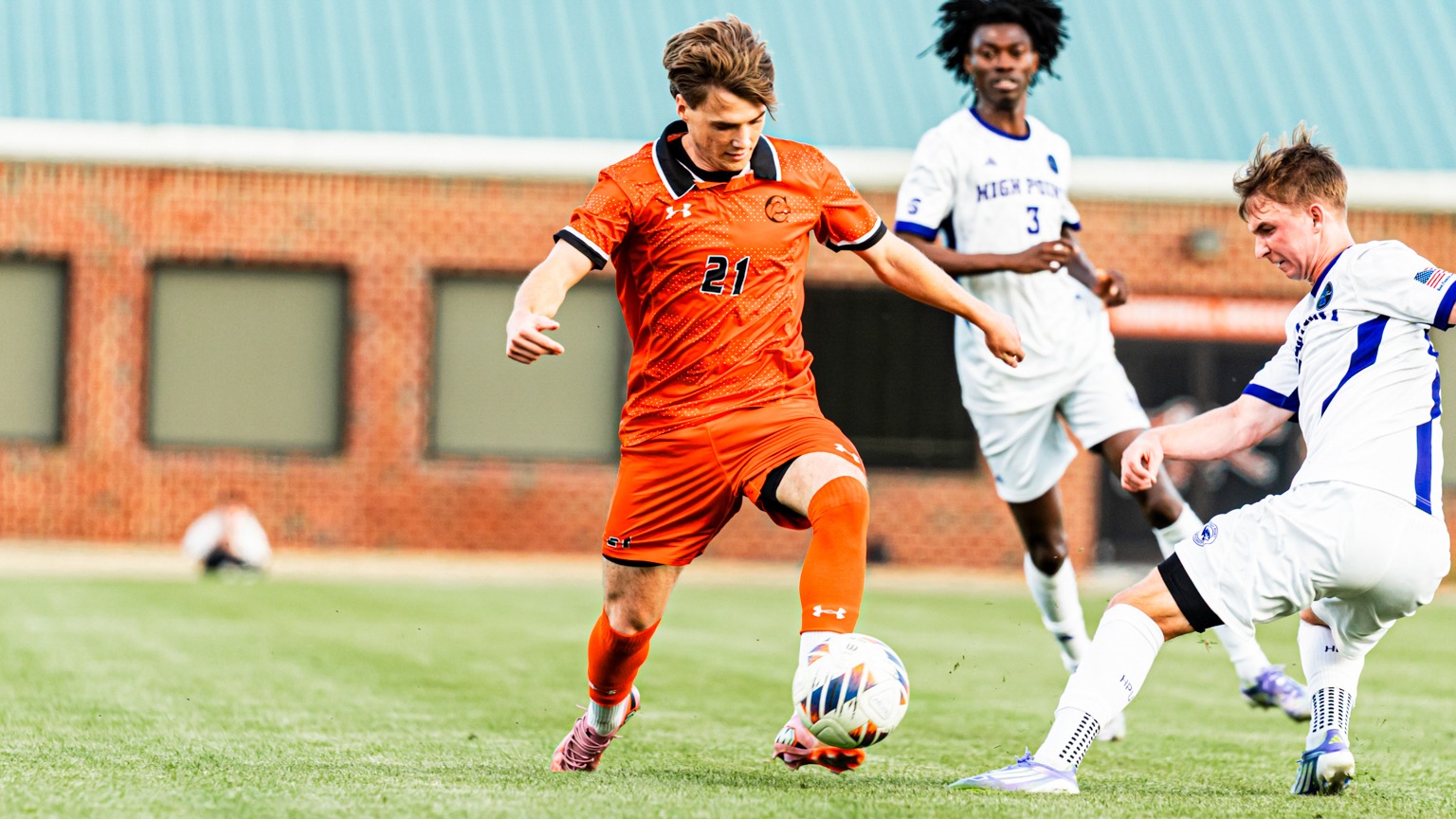 Xander Martin dribbling a soccer ball around a player from High Point University with another HPU player in the background