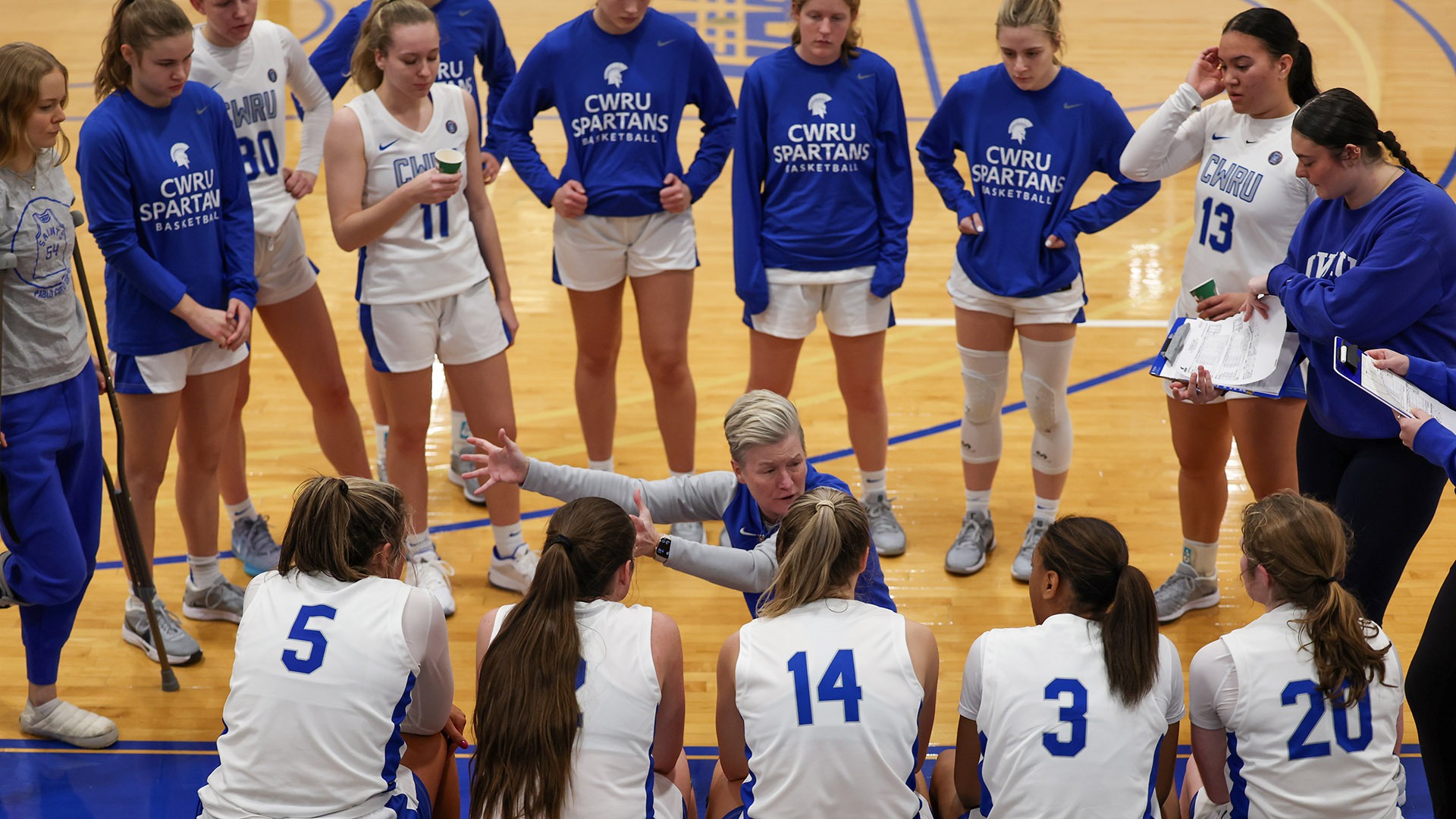 The CWRU women's basketball team in a circle on the bench listening to Coach Reimer during a timeout against Carnegie Mellon