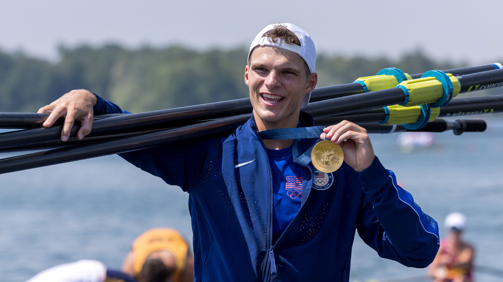 Cornell men's heavyweight rowing alum Michael Grady '19 carries oars across his shoulders and holds his gold medal in his right hand after claiming first place in the men's four at the Vaires-sur-Marne Nautical Stadium on Thursday, Aug. 1, 2024, at the 2024 Olympics.