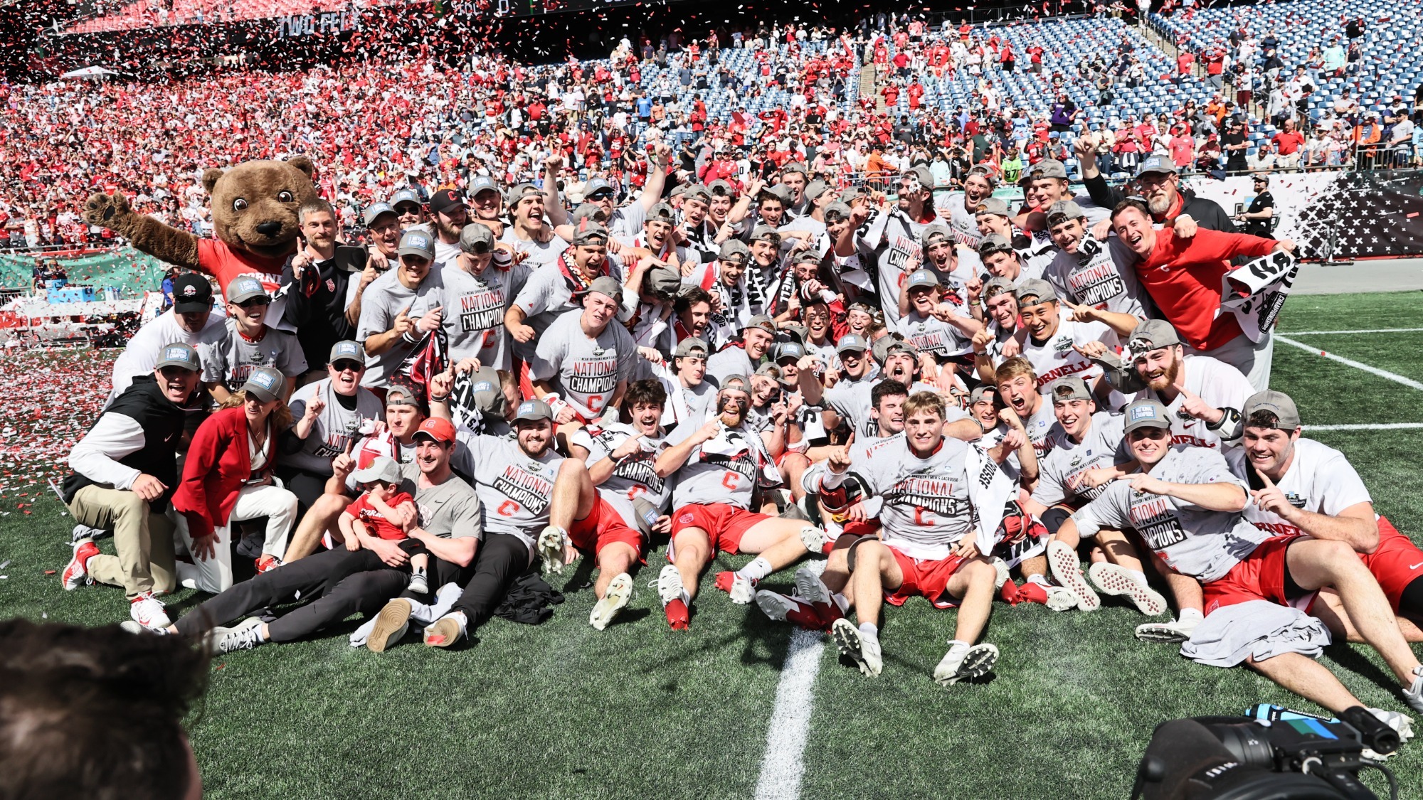 The Cornell men's lacrosse team celebrates the 2025 NCAA championship on the field at Gilette Stadium with a team picture with confetti flying in the air.